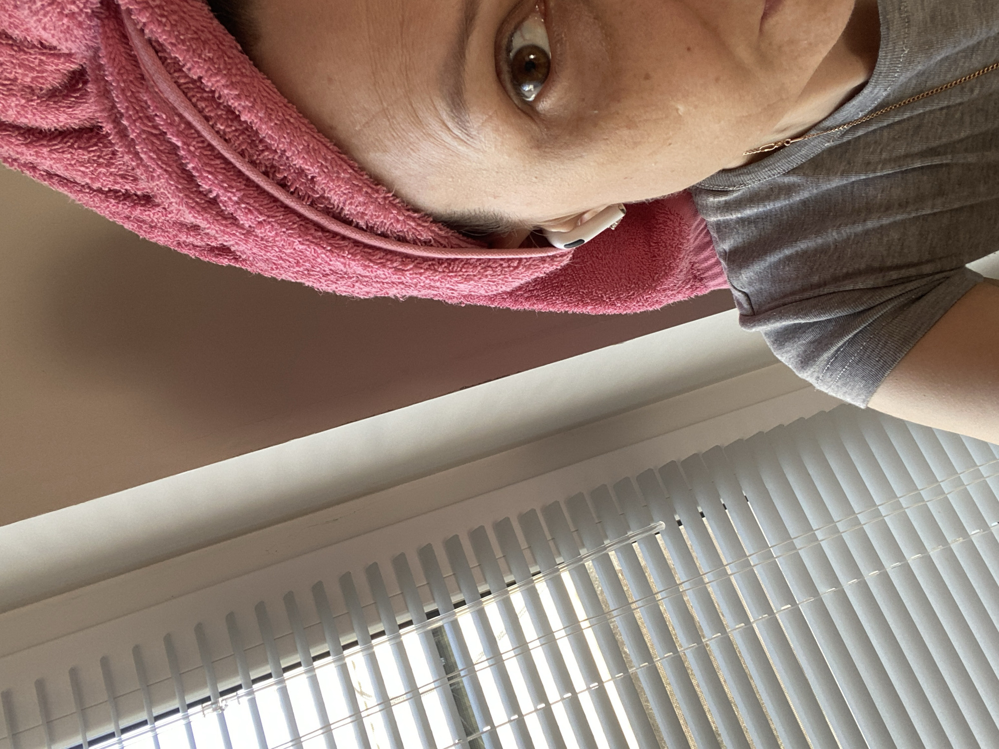 The scene depicts a close-up indoor view from Chaikovsky, Russia. The right side of an adult female's face is visible, featuring a brown eye looking directly towards the viewer. Her hair is wrapped in a pink terrycloth towel in a turban style. A white wireless earbud is present in her right ear. She is wearing a grey short-sleeved t-shirt, and a thin gold-colored chain is visible around her neck. To the left, a window fitted with white horizontal Venetian blinds allows diffused natural light into the space. A light beige wall is visible in the upper right background.