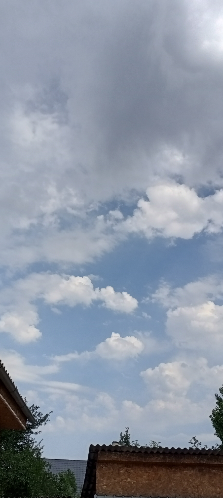 The image captures a view of the sky with scattered clouds, typical of a partly cloudy day. In the foreground, the roofs of two structures are visible, one with a lighter colored, wooden facade and a corrugated roof, and another on the left with a darker, possibly tiled roof and a wooden overhang. Green foliage from trees and bushes is present in the lower portions of the frame, peeking from behind and alongside the structures. The sky occupies the majority of the image, with areas of bright blue interspersed with various forms of white and gray clouds. The overall impression is of an outdoor scene, likely in a residential or semi-rural area. There are no people or animals visible in the image.