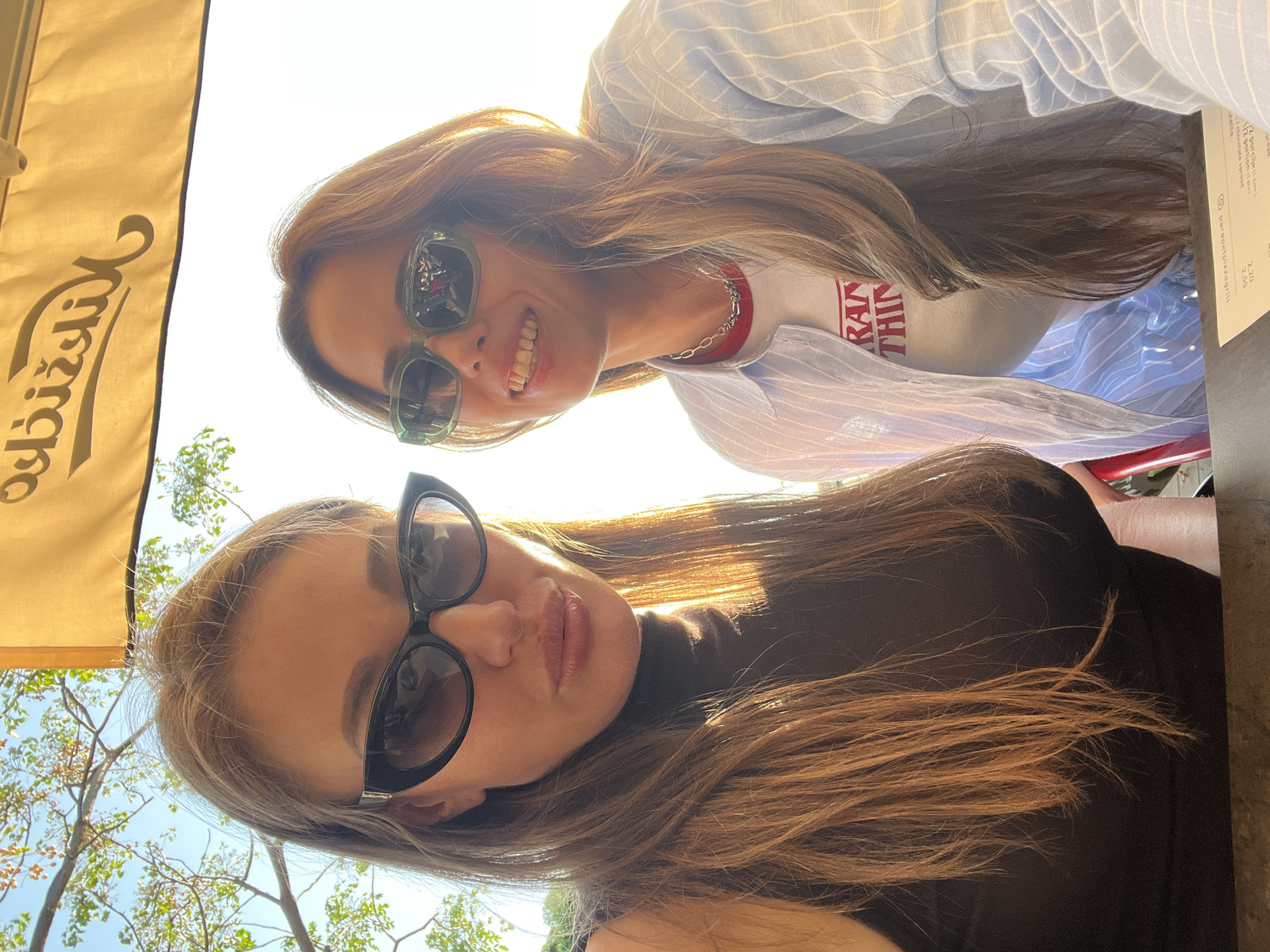 Two women are looking directly at the camera. The first woman is wearing a black top and black sunglasses. The second woman is wearing sunglasses with green frames, a light blue striped shirt, and a white t-shirt with the words "STRANGER THINGS" written on it. Both women have long brown hair. A menu card is partially visible on the upper right side of the image. The image appears to have been taken under a yellow awning, possibly at a restaurant or cafe. Trees and a clear sky are visible. The location is Bar, Montenegro.