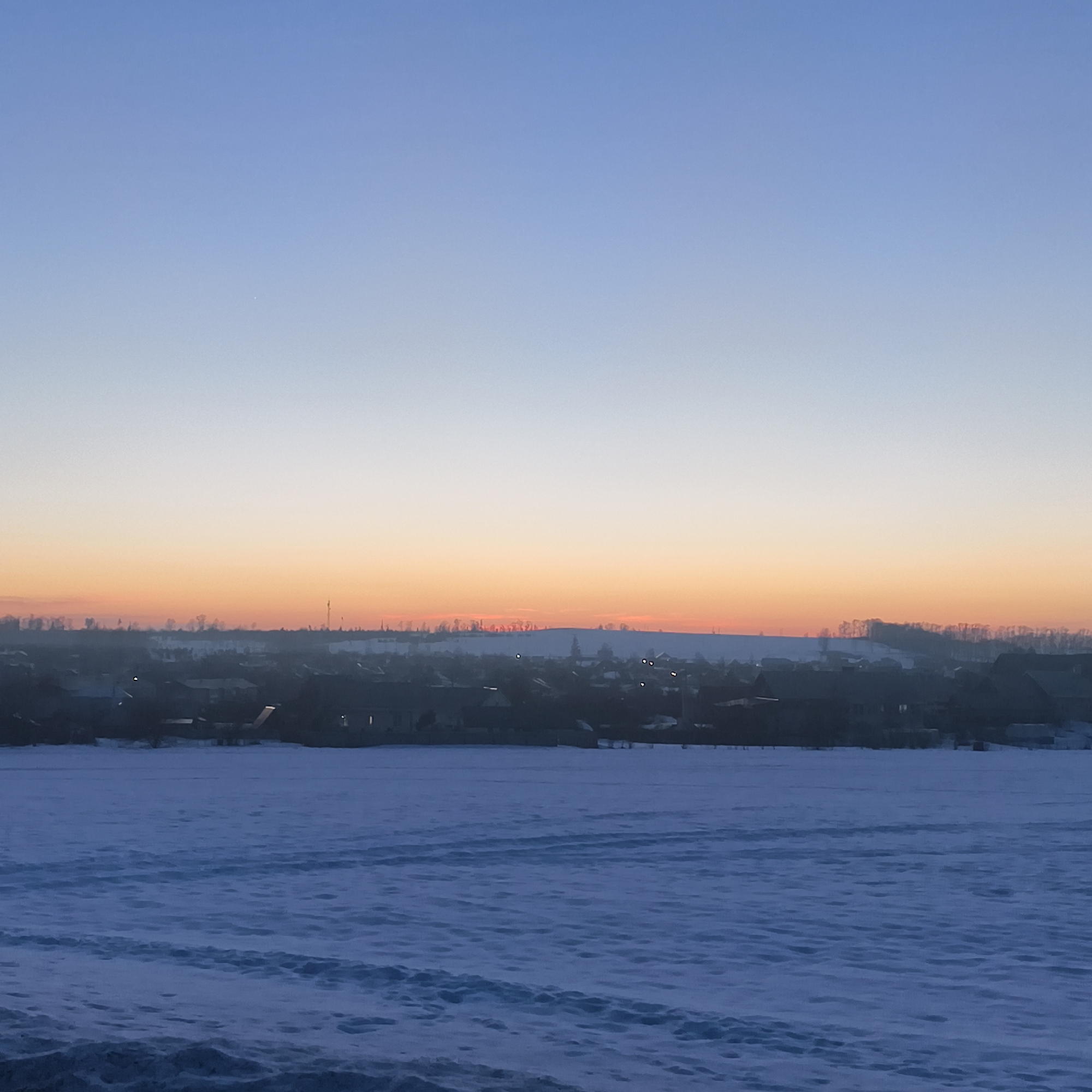 The image shows a winter landscape in Фоки, Russia. In the foreground is a snow-covered field with visible tracks. In the middle ground, a dark silhouette of a village or town is visible. The sky is a gradient of color, transitioning from blue at the top to orange near the horizon, indicating sunrise or sunset. No people are visible.