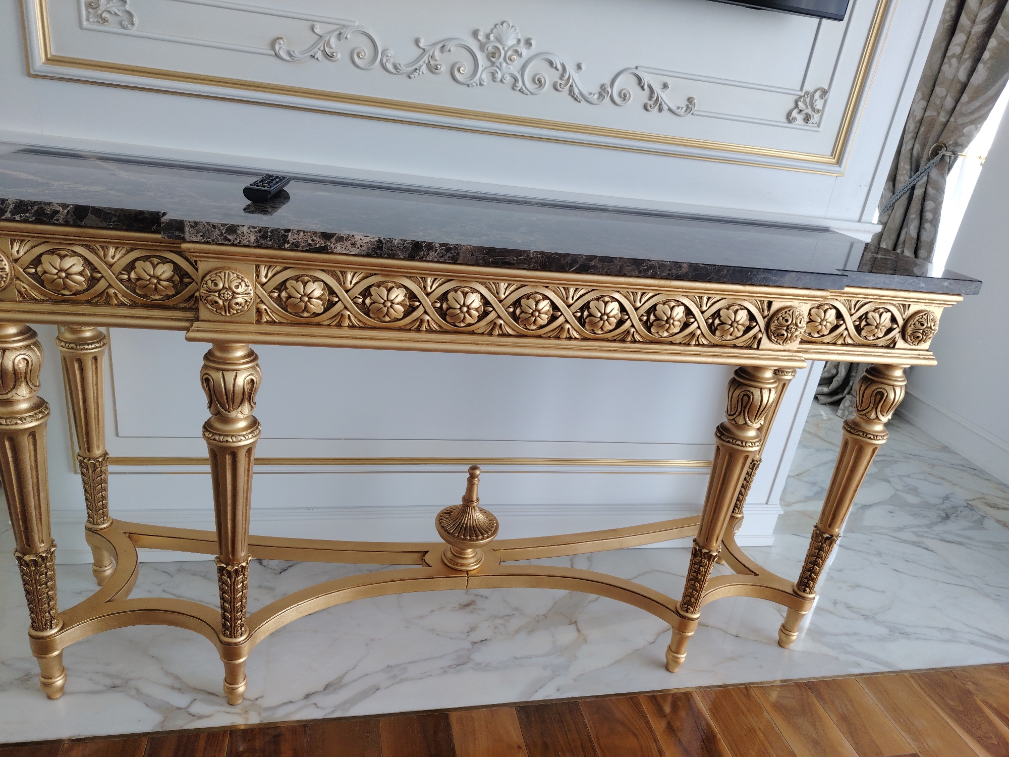 The image shows a close-up, eye-level, full shot of an ornate, gold-colored console table with a marble top. The table is placed against a white wall with elaborate gold trim and decorative molding. A black remote control rests on the marble surface of the table. The floor beneath the table is a mix of white marble and wooden planks. The overall style of the room appears to be luxurious and classical. The location is identified as Konigil, Uzbekistan.