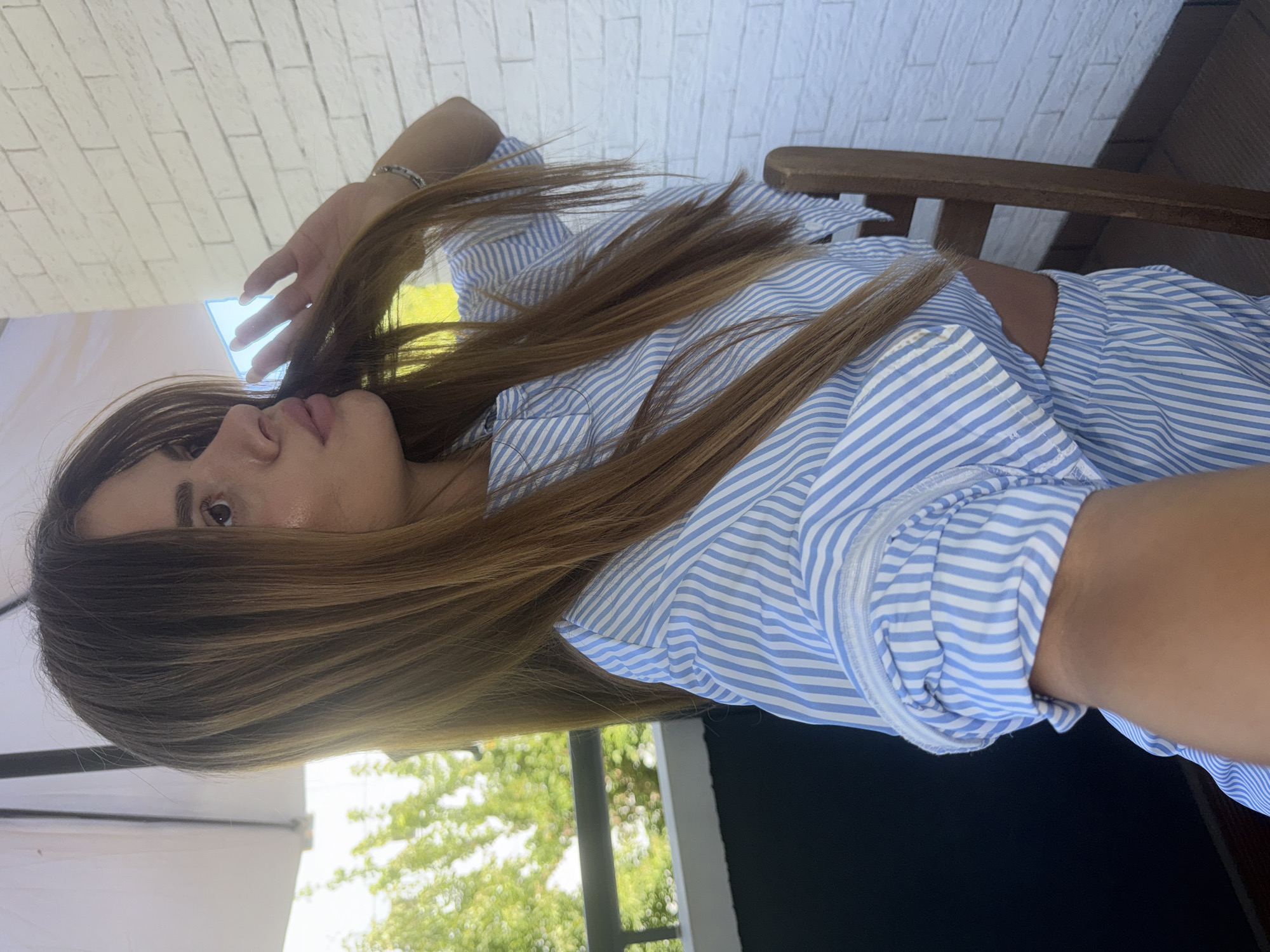 The image shows a young woman with long, brown hair, wearing a blue and white striped crop top and matching shorts. She is sitting outdoors, on what appears to be a balcony or patio. In the background, there is a white brick wall and some greenery from trees. A wooden chair is visible to the right of the woman. The scene suggests a casual, sunny day. The location is the city of Plovdiv, Bulgaria.