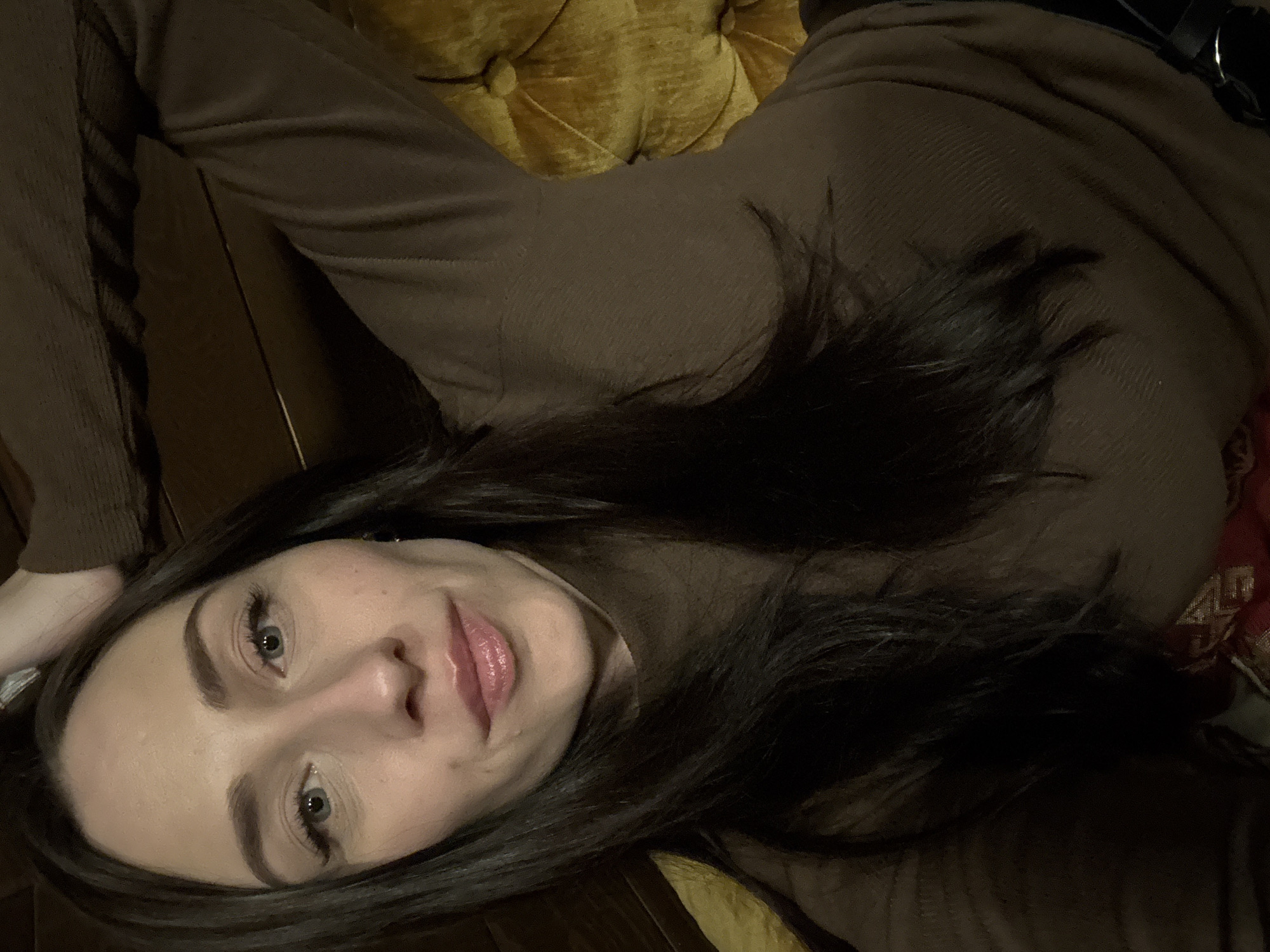 A woman is lying on a brown upholstered surface with tufted buttons, possibly a sofa or chaise lounge. She is wearing a long-sleeved brown top. Her head is positioned toward the bottom of the frame, with her dark hair partially obscuring her shoulder. The background is dark, but wooden floor is visible. The setting appears to be indoors. There is no immediate indication that the image was taken in Saint Petersburg, Russia.