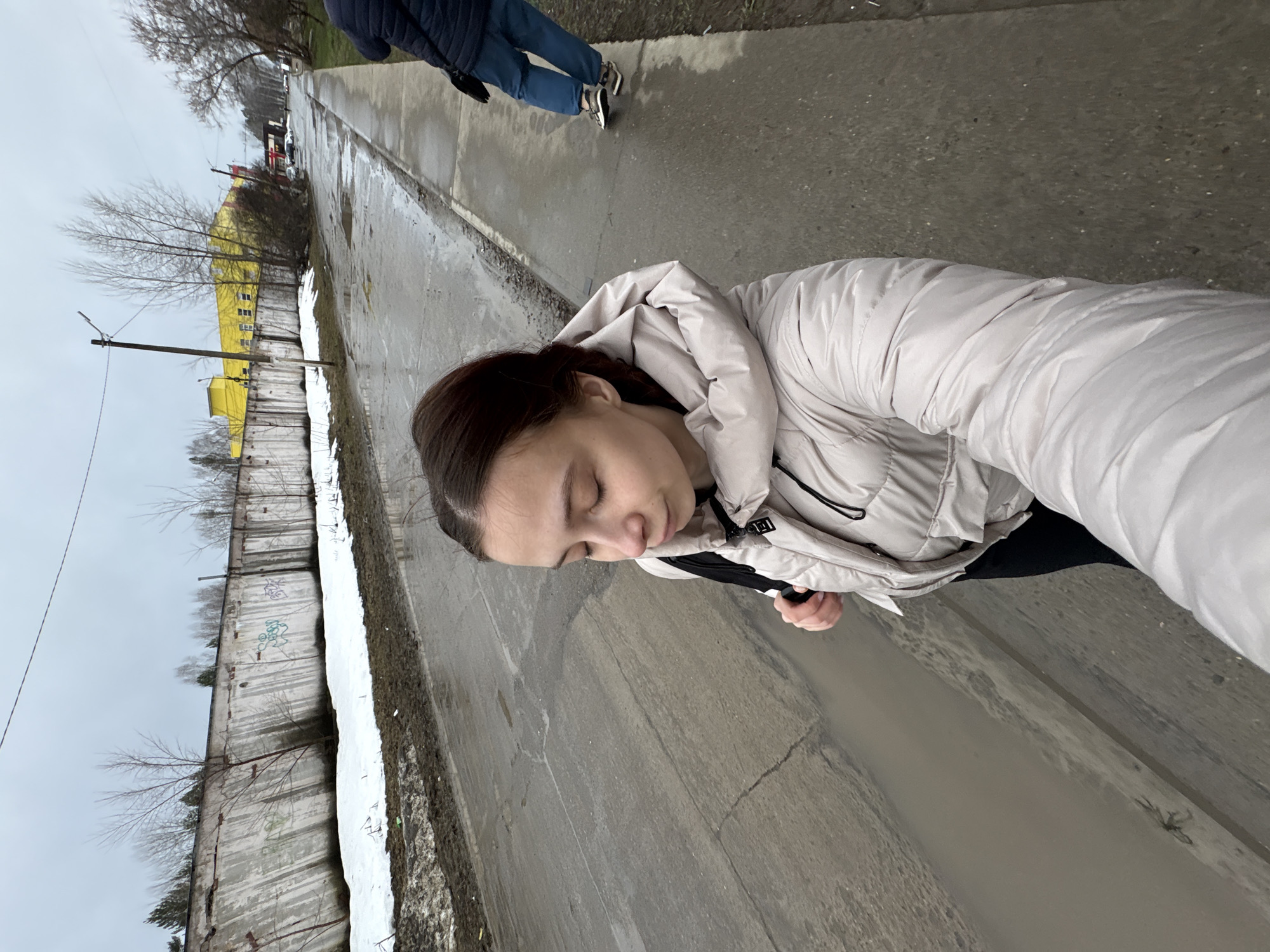 A woman in a light-colored puffer jacket stands on a wet sidewalk. Her eyes are closed, and her head is tilted slightly downwards. Another person's legs are visible in the upper part of the frame, indicating they are walking nearby. The sidewalk is next to a long, concrete wall. A tall, yellow building is visible in the background. The sky is overcast. The location is Chaikovsky, Russia.