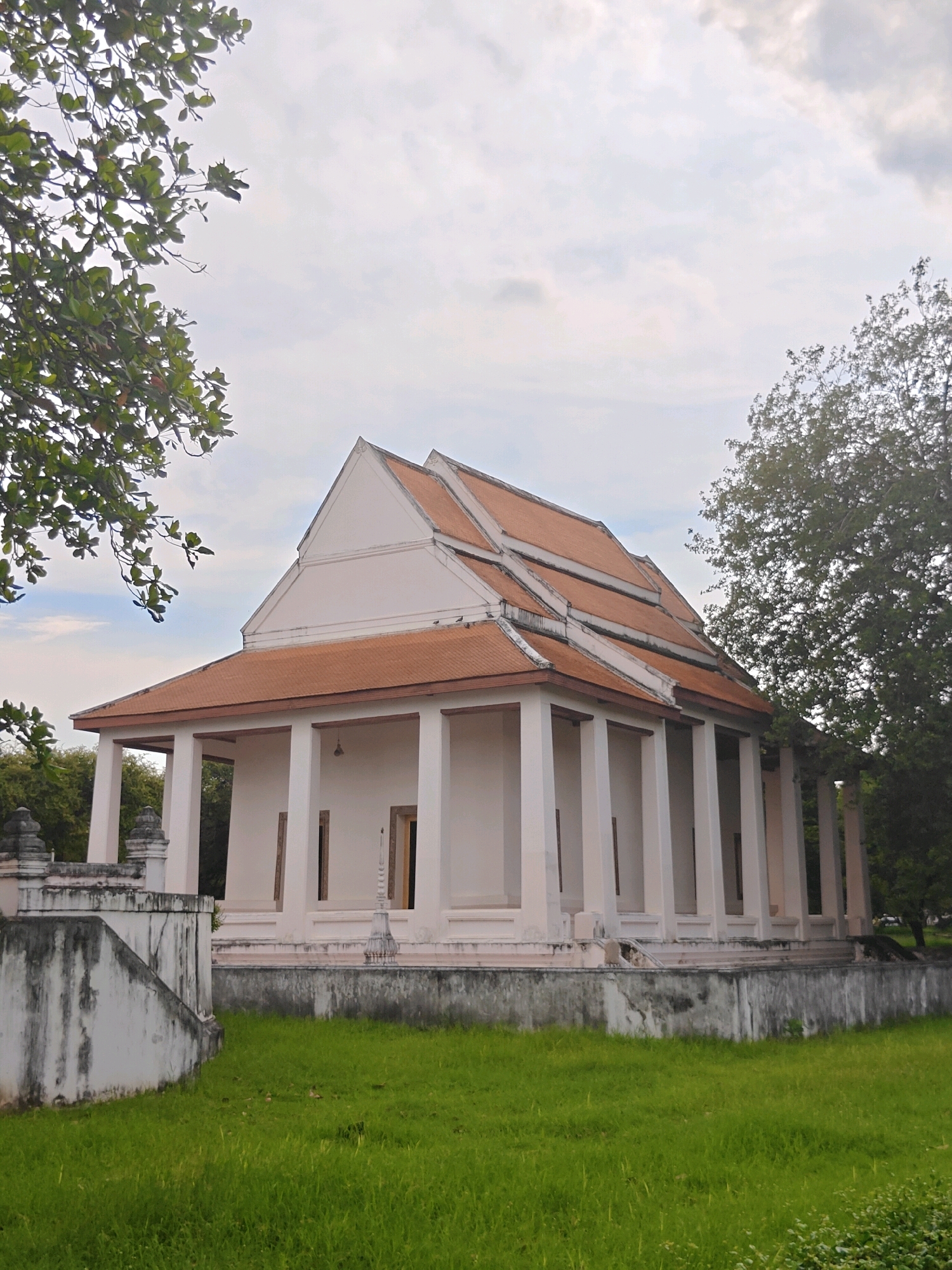 The image shows a white building with an orange tiled roof, supported by several white pillars. The building appears to be a temple or a historical structure. It is surrounded by lush green grass and trees. In the foreground, a grey concrete structure with a staircase leads up to the building. The sky is overcast with clouds. The style of the architecture suggests it is located in Southeast Asia, possibly Thailand. Based on the architectural style, this building is likely located in Phra Nakhon Si Ayutthaya City Municipality, Thailand, a region known for its ancient temples and historical ruins. There are no people or animals visible in the image.