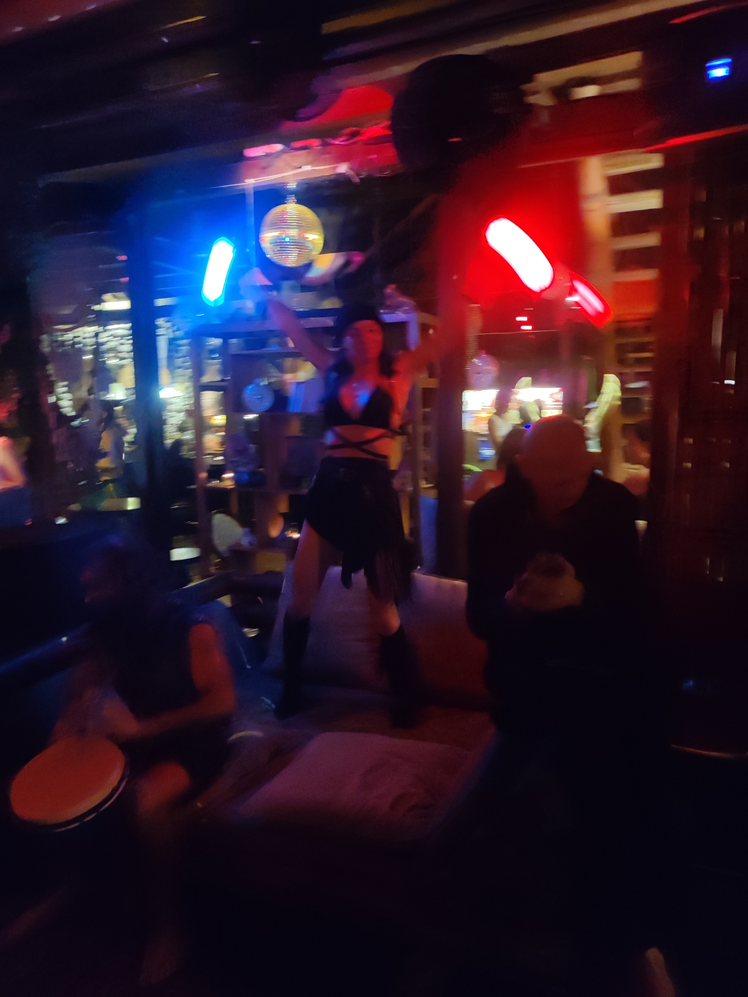 The scene is set in a dimly lit indoor space with a vibrant atmosphere. The location may be a bar or nightclub in Lokaserena, Indonesia, suggested by the overall vibe. A woman in a dark outfit, possibly a dancer, stands with arms raised, center frame. To her left, a man is seated, playing a hand drum. To her right, another person sits, mostly obscured by shadows. The background shows indistinct figures and lights, indicating other patrons and the location's decorative elements. There's a disco ball and colored lights contribute to the atmosphere.