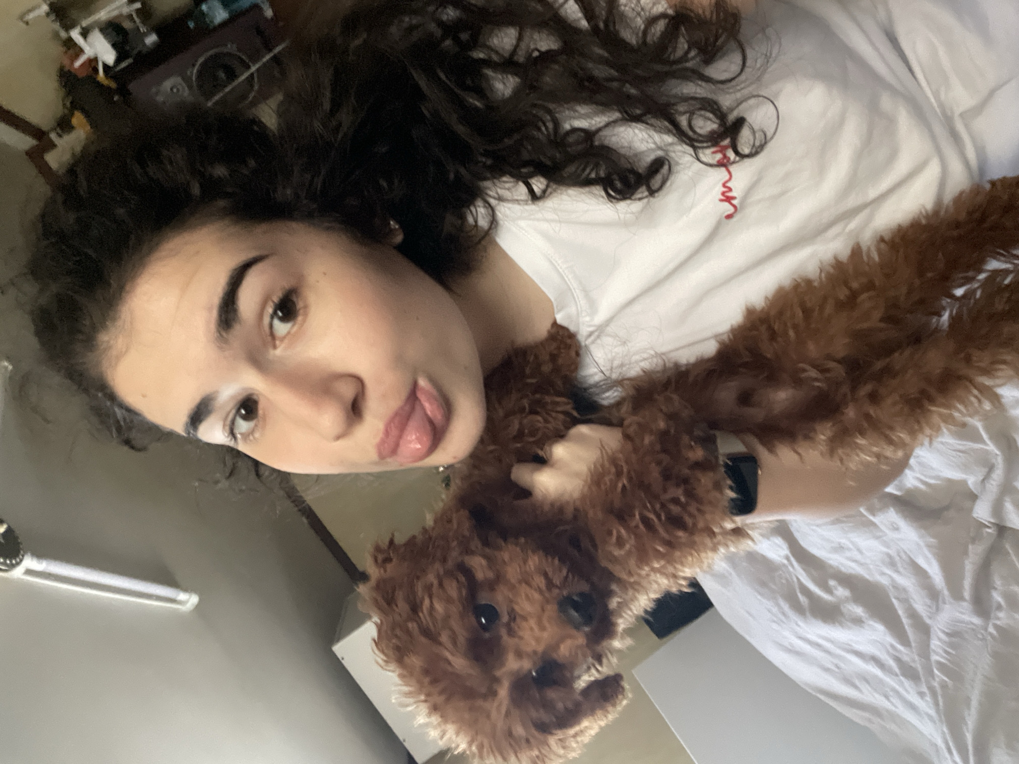 A young woman with curly dark hair and light skin is holding a brown poodle. She is sticking her tongue out at the camera. She is wearing a white T-shirt and a black smart watch. The location is indoors, possibly a bedroom, and the setting is casual. The location is not specified.