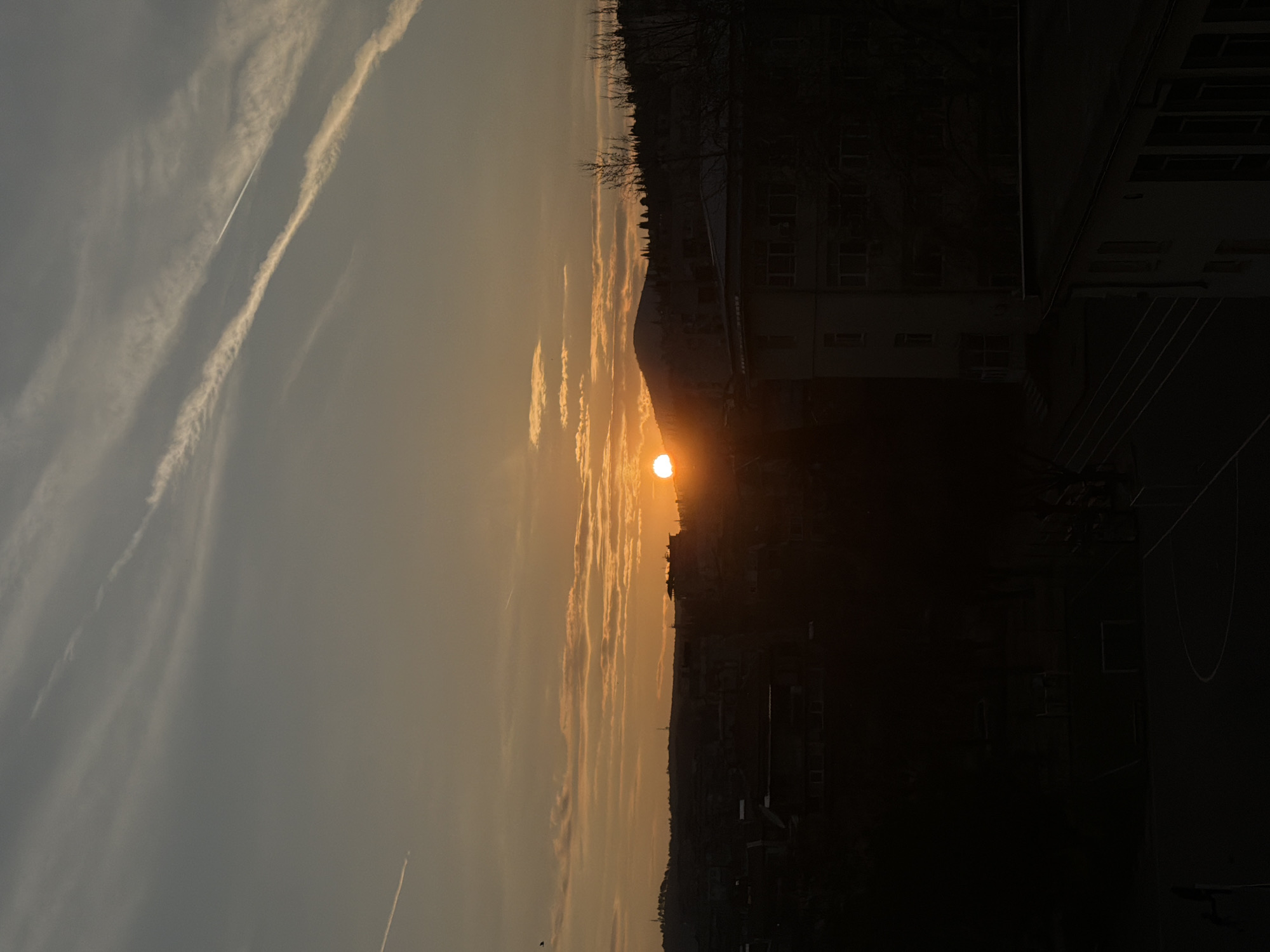 The image displays an urban landscape in Stara Zagora, Bulgaria, at sunset. The sky transitions from a cool grey-blue at the top to a warm orange-yellow near the horizon, exhibiting several horizontal cloud formations. Two prominent, parallel contrails are visible in the upper right quadrant, with a fainter contrail in the upper left. A small, dark, winged object is discernible in the upper left. The sun, a bright orange-yellow orb, is partially obscured by a distant, silhouetted hill or mountain range positioned slightly right of the frame's horizontal center.
The mid-ground features the silhouette of the city, with a prominent range of hills or mountains extending across the horizon, topped with sparse, bare trees and several communication towers. A multi-story building with visible window structures is present on the right side of the frame, largely in shadow. The immediate foreground is enveloped in deep shadow, faintly revealing the markings of a sports court, including what appears to be a basketball hoop and a soccer goal, outlined by white lines on a dark surface. No individuals or active interactions are visible.
