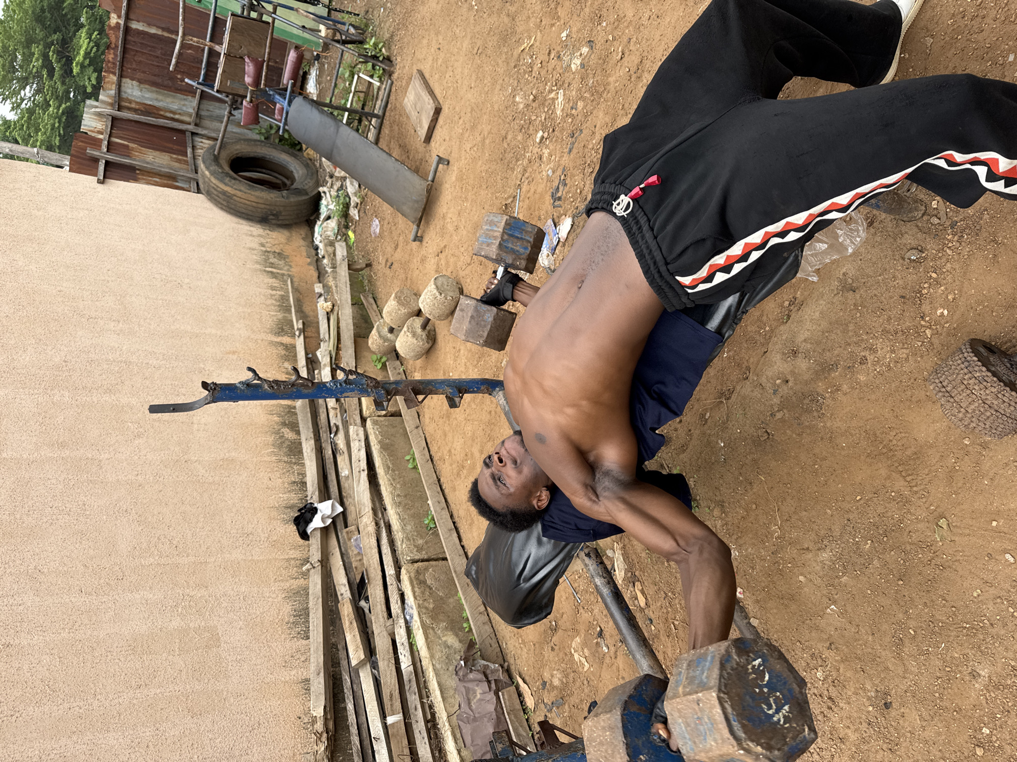In Akwanga, Nigeria, a young Black man is performing a dumbbell chest press on a weight bench outdoors. He is lying on his back on a black padded bench, with his arms extended upwards, holding a large, hexagonal dumbbell in each hand. The dumbbells are made of metal and appear to be heavy. He is wearing black sweatpants with a red, white, and black stripe down the side of each leg, and a dark blue t-shirt. His face is contorted in an expression of exertion.
The surrounding environment is a rough, outdoor training area. The ground is a mix of dirt and gravel. In the background, there is a textured, light-colored wall, piles of wooden planks, some large concrete blocks, a rusty metal fence, and a worn-out tire. There is also a metal weight rack with a faded green seat and a rusted metal incline bench in the background. The overall impression is of a makeshift outdoor gym.