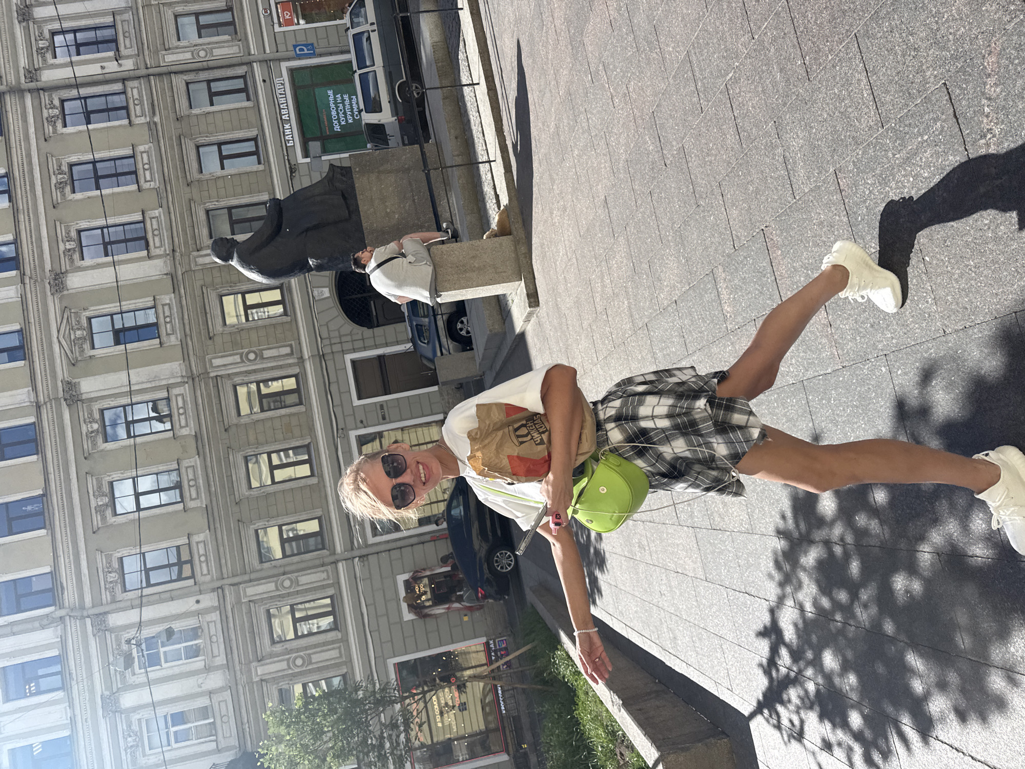 The image depicts a woman walking on a sunlit, paved city street in Saint Petersburg, Russia. She is smiling and looking towards the camera, holding a paper bag in her right hand and a mobile phone in her left. She is wearing a white t-shirt, a black and white plaid skirt, sunglasses, and white sneakers. A bright green purse is slung over her left shoulder. In the background, there is a grand, old building with many windows, characteristic of St. Petersburg architecture. A statue of a seated man is visible in the middle ground, along with a bank sign in Russian that reads "БАНК АВАНГАРД". A man is sitting on a pedestal near the statue, and a cat is lying at the base. The street is lined with trees, and shadows from the leaves are cast on the pavement. Cars are parked and driving on the street. The overall scene suggests a typical summer day in the city.
