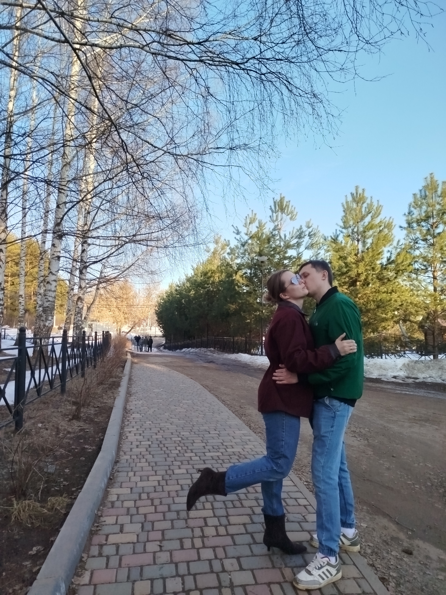 The image depicts a couple kissing on a brick pathway in Izhevsk, Russia. The woman has her right leg raised and is wearing sunglasses, a burgundy jacket, blue jeans, and black boots. The man is wearing a green jacket, blue jeans, and white sneakers with grey stripes. They are embracing.
To their left is a black metal fence along the pathway, lined with bare trees. In the background are evergreen trees and a group of people walking in the distance. To their right, the path turns into dirt. The sky is blue.