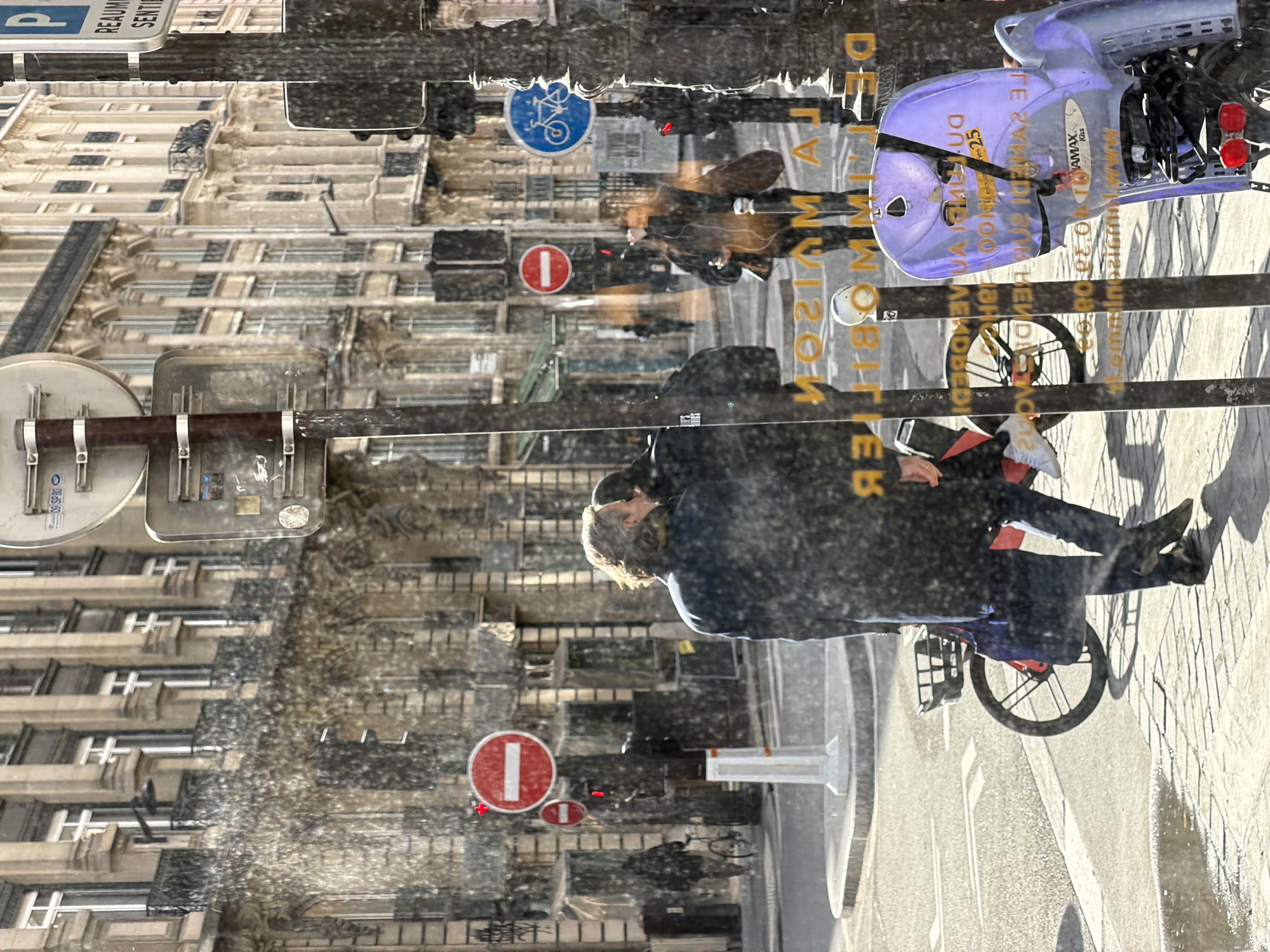 The image depicts a reflection in a window, capturing a street scene in Paris, France. In the reflection, a person is riding a bicycle along a street with light-colored paving stones. The bicycle appears to be a rental bike. Reflections of Parisian architecture are visible in the window, including multi-story buildings with windows and ornate facades. Road signs are reflected as well, including a "no entry" sign (a red circle with a white horizontal bar) and a sign indicating a bicycle route. The reflections create a layered and somewhat distorted view of the scene, blending the street activity with the city backdrop.