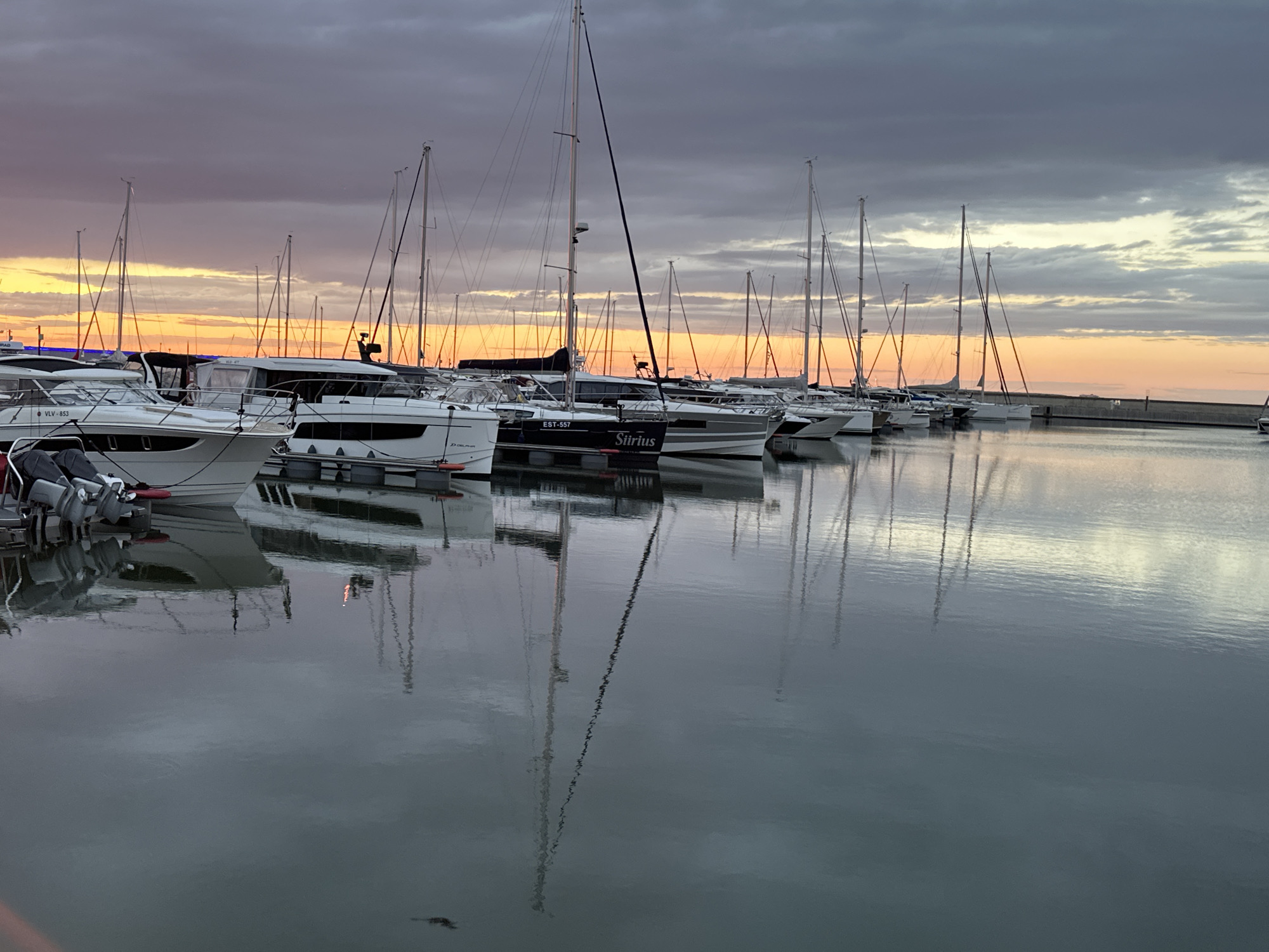 The image depicts a marina in Tallinn, Estonia, at dusk. A fleet of sailboats and motorboats are docked, their masts creating a striking silhouette against a dramatic sky. The sky is a gradient of deep gray clouds transitioning to vibrant orange and yellow hues near the horizon, indicative of a sunset or sunrise. The calm water of the marina reflects the boats and the colorful sky, creating a mirror-like effect. Several boats are visible in the foreground and middle ground, with details like names such as "Sirius" and registration numbers like "EST-557" on some of them. The overall scene conveys a sense of tranquility and natural beauty, common in coastal cities during twilight hours.