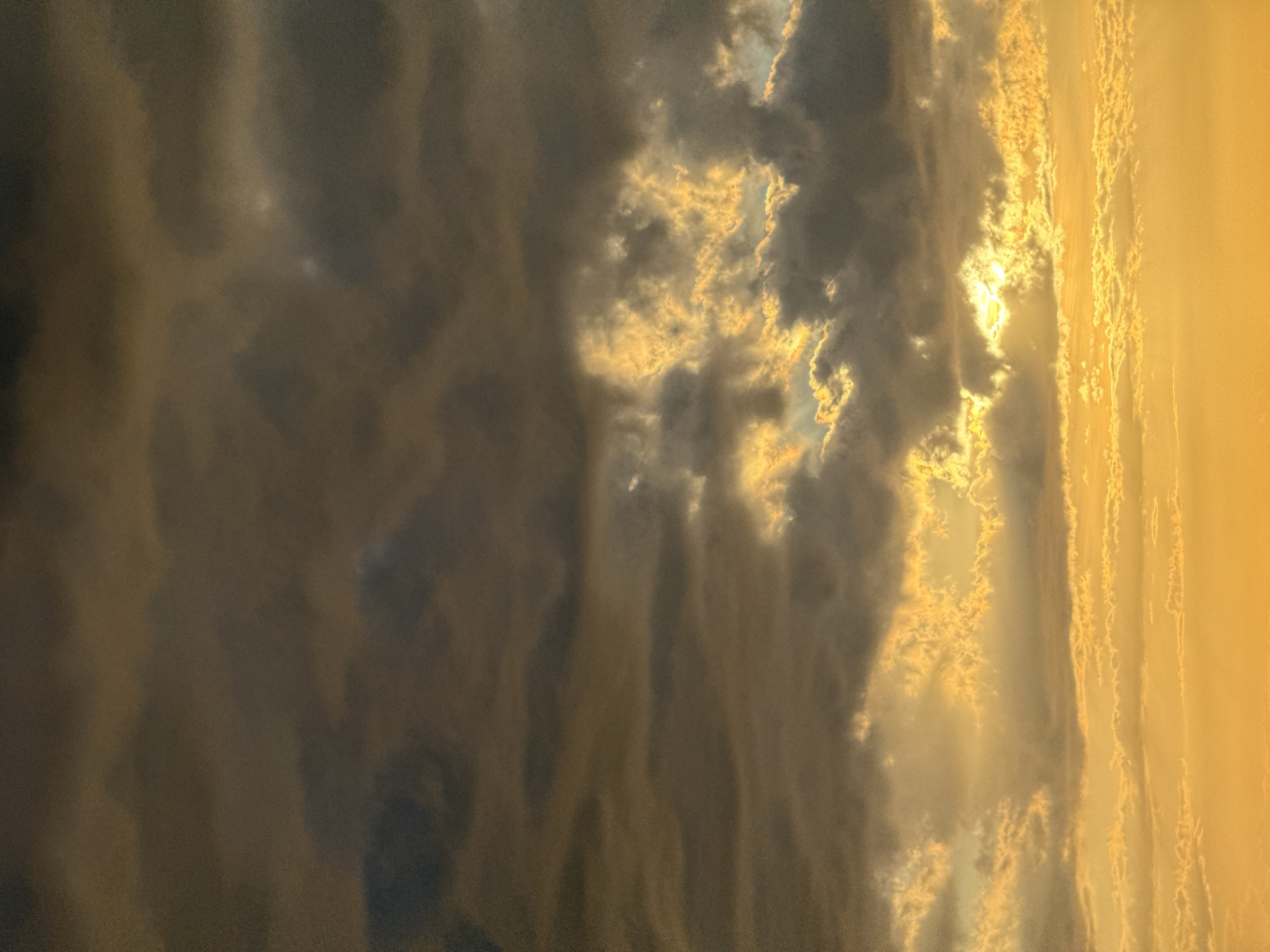 This image captures a dramatic sky scene with dense, dark clouds interspersed with bright, golden light. The clouds appear textured and voluminous, with some illuminated from behind, suggesting a sunset or sunrise. The overall impression is one of a powerful atmospheric display.
The scene is dominated by the sky, with no discernible objects or people. The composition is vertical, emphasizing the scale and texture of the clouds. The lighting creates a strong contrast between the dark, shadowed areas of the clouds and the radiant, golden hues that break through. These illuminated sections suggest breaks in the cloud cover, allowing sunlight to pass.
The image evokes a sense of awe and the sublime, characteristic of dramatic weather phenomena. The golden light can be interpreted as hopeful or majestic, while the dark clouds add a sense of drama and power. Given the location context of Baan Plai Laem, Thailand, this could be a view of the sky over a tropical landscape, potentially near the coast. The time of day is clearly indicated by the warm, golden light, pointing towards either early morning or late evening. The weather appears to be dynamic, with a mix of heavy cloud cover and clear, sunlit breaks.
There is no visible text in the image.