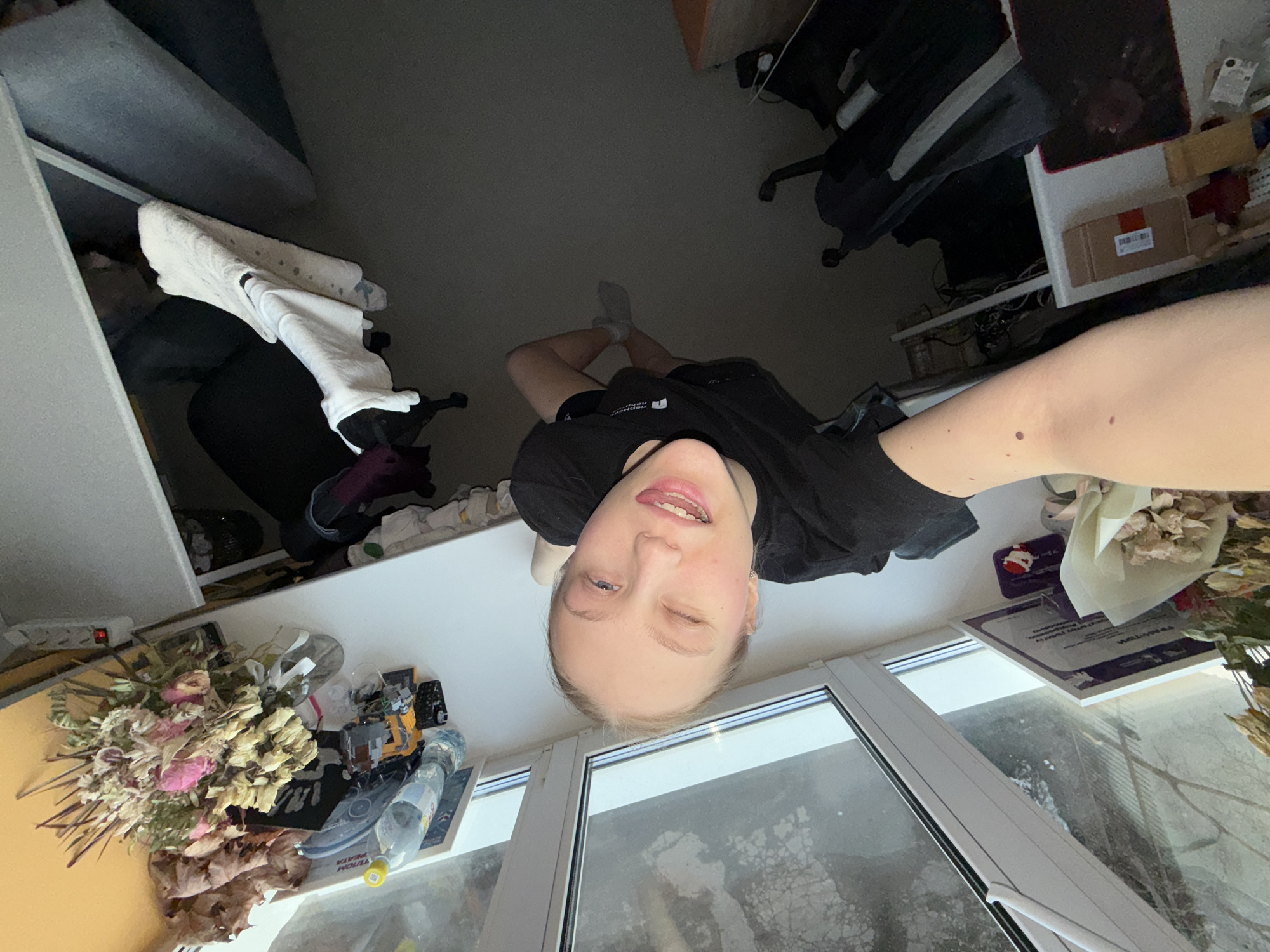 A woman is lying upside down on a window sill, looking directly at the camera. She is wearing a black t-shirt and her eyes are open. Outside the window, there is a view of a cityscape. On the window sill, there are dried flowers, a bottle of water, and other objects. To the left of the woman is a bed and to the right is a desk. The location is likely in Perm, Russia.