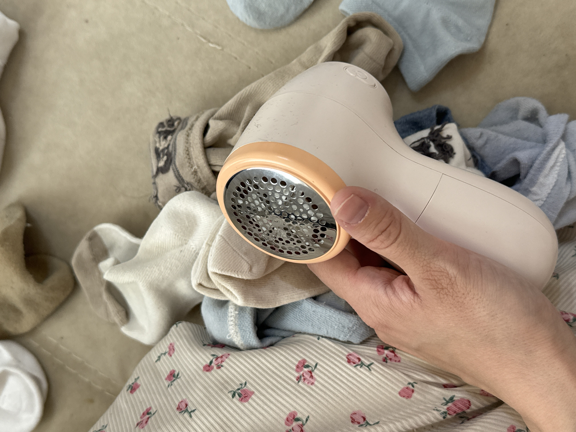 A person's right hand holds a peach-colored electric lint shaver over a pile of socks and a beige and white striped shirt with pink rose patterns. The background is a light beige surface. The location is unspecified in the image and cannot be inferred to be Chaikovsky, Russia.