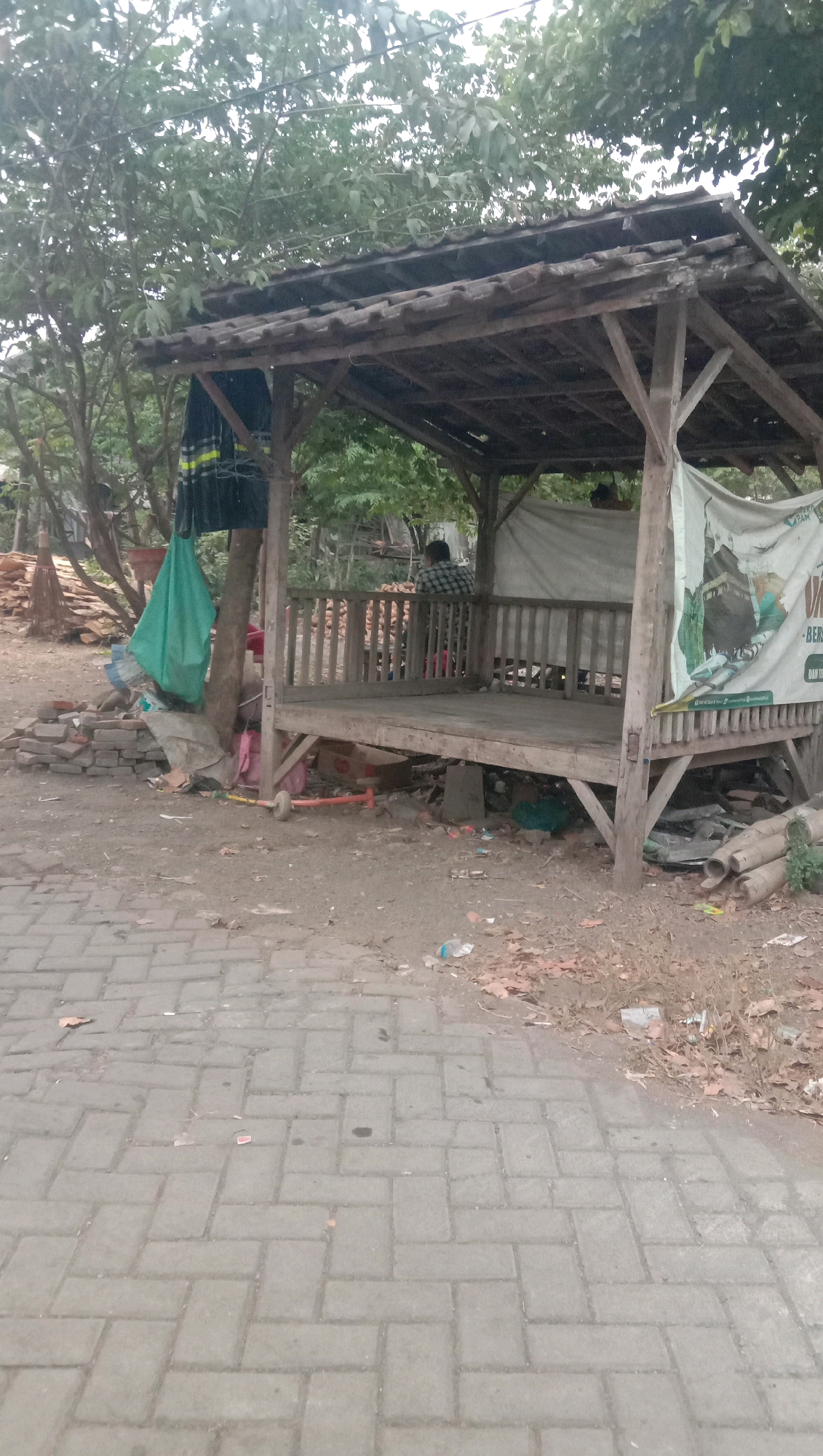 A rustic wooden pavilion sits on a dirt and paved ground area. The pavilion has a weathered tile roof supported by wooden posts and beams. It features a wooden floor and a railing around the perimeter. Inside, a person wearing a plaid shirt is seated, facing away from the camera. To the left of the pavilion, a large green tarp is hung from a tree, along with other assorted items. Piles of bricks and debris are scattered around the base of the pavilion and the surrounding area. A paved walkway with interlocking bricks extends across the foreground. In the background, trees and foliage create a natural backdrop. A banner with graphics and text is attached to the right side of the pavilion. The scene suggests an outdoor, possibly semi-abandoned, public or private space.