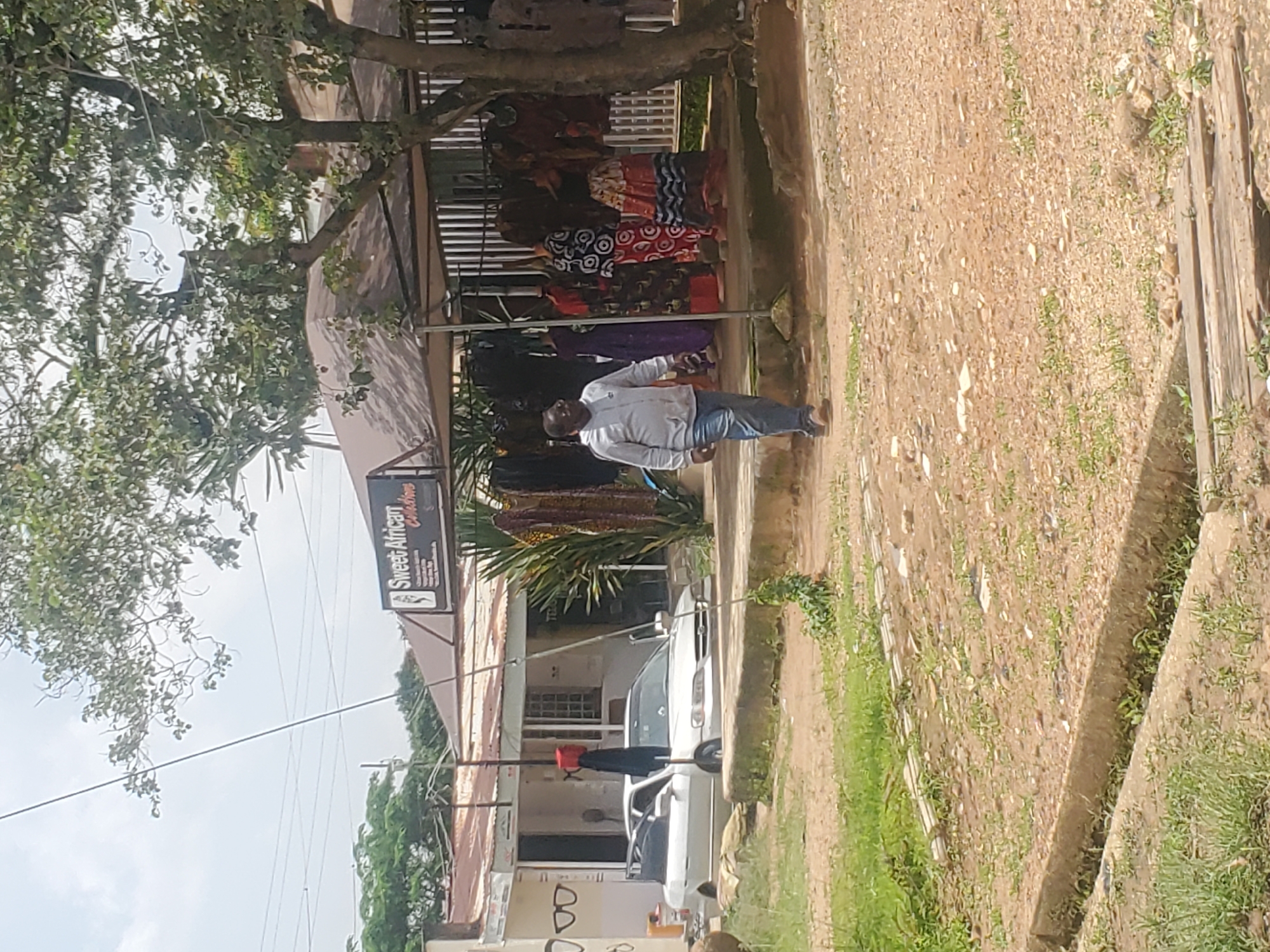 The image depicts a street scene in Jos, Nigeria, possibly near a market or commercial area. In the foreground, a dirt path with a drainage ditch is visible, bordered by grass and some scattered debris. A wooden plank bridge crosses the ditch.
In the mid-ground, a silver sedan car with its doors open is parked on a paved surface in front of a building. To the right of the car, a stall or shop is set up under a covered structure. This stall displays a variety of colorful African-print clothing on mannequins and hanging racks. A sign above the stall reads "Sweet African Collections."
A man in a light gray hooded sweatshirt and blue jeans is walking from left to right in front of the stall. He is wearing glasses and appears to be looking towards the stall. Several trees with green foliage are present, partially obscuring the sky and the buildings. The overall impression is a typical street view of a business district in Jos.