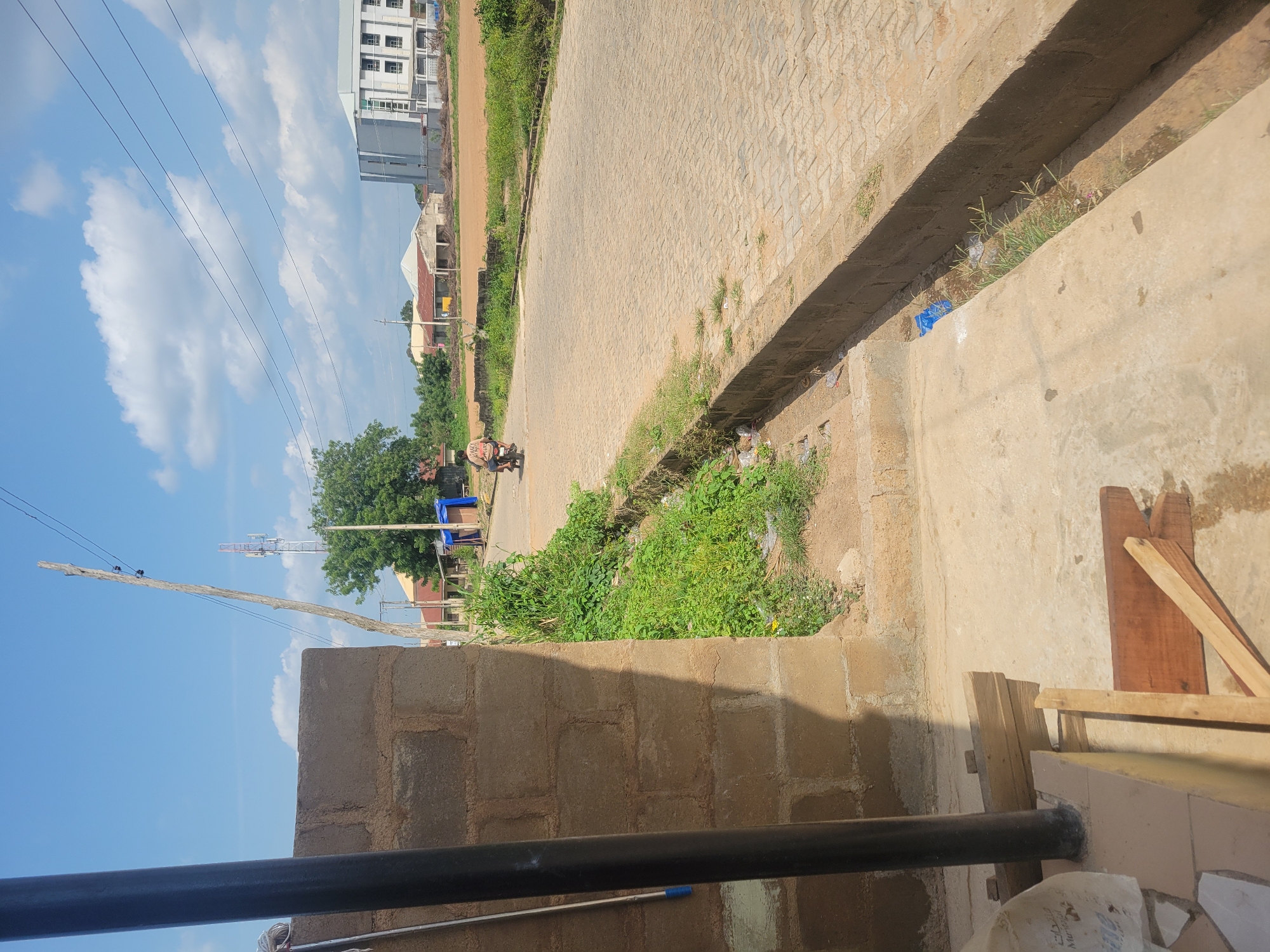 The image depicts a street scene in Ogbomosho, Nigeria, on a sunny day with a clear blue sky and scattered white clouds. The perspective is from ground level, looking slightly upwards. A paved road, constructed from interlocking bricks, dominates the mid-ground, running diagonally across the frame. A concrete drainage channel runs alongside the road on the right, filled with some vegetation and debris.
In the background, a multi-story building with a light-colored facade and numerous windows stands on the left. Adjacent to it, there are other buildings with tin roofs. A large, leafy green tree is visible further down the street. Power lines crisscross the sky, supported by a wooden pole in the foreground and a metal tower in the distance.
In the immediate foreground, a section of a brick wall and a dark metal pipe are visible. A mop handle rests against the wall. On the road, a person is riding a motorcycle, their back to the camera. The overall impression is of a typical urban or semi-urban environment in Nigeria during daylight. No visible text is present in the image.