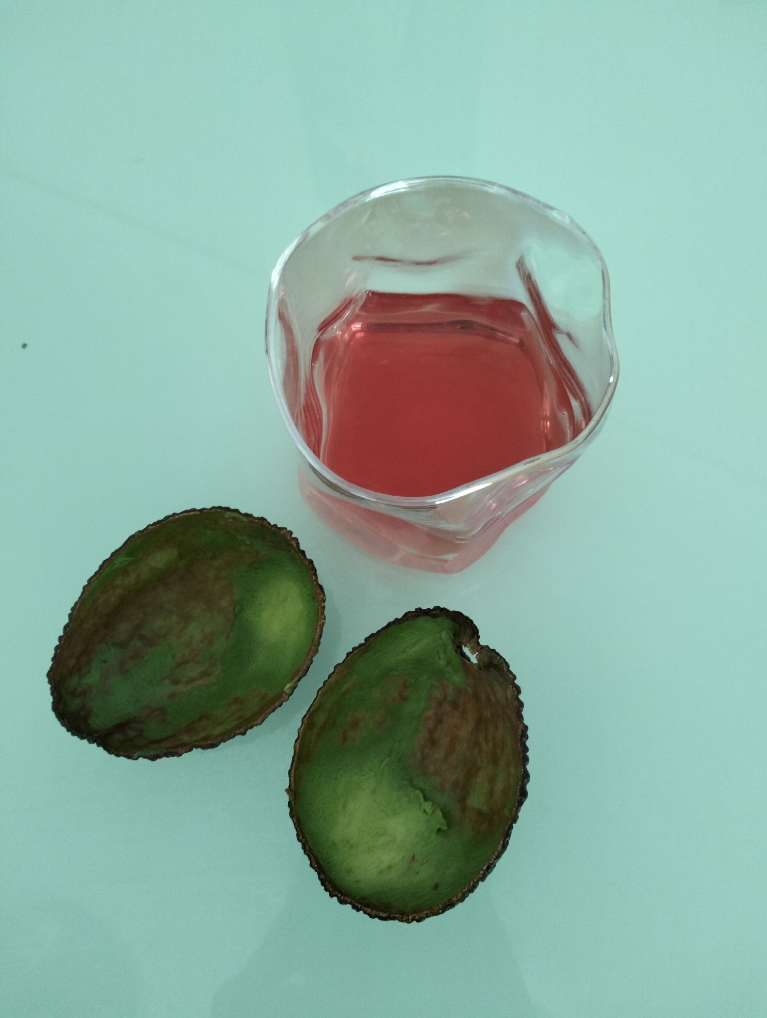 A glass of red liquid is positioned near two avocado halves on a light blue surface. The scene may be located in Krykhivtsi, Ukraine.