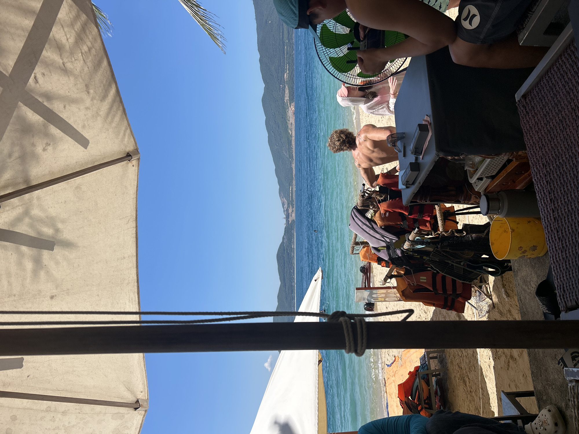 The image depicts a sunny day at a beach in Bac Nha Trang Ward, Vietnam, viewed from under a beach umbrella. In the foreground, a dark pole with rope is visible. To the right, a person's arm and shoulder are in frame, holding a remote control. A large, green fan is positioned in front of them. Behind the fan, a man with a muscular back and blonde, curly hair stands facing away from the camera, looking towards the ocean. Next to him, a person wearing a white hooded garment and a pink cap is also facing away. In the middle ground, a rack holds several orange life vests and other items. The beach is sandy, with a few scattered items including a yellow bucket and a white umbrella in the background. The ocean is a vibrant turquoise, stretching to the horizon where rolling green hills form the backdrop under a clear blue sky.