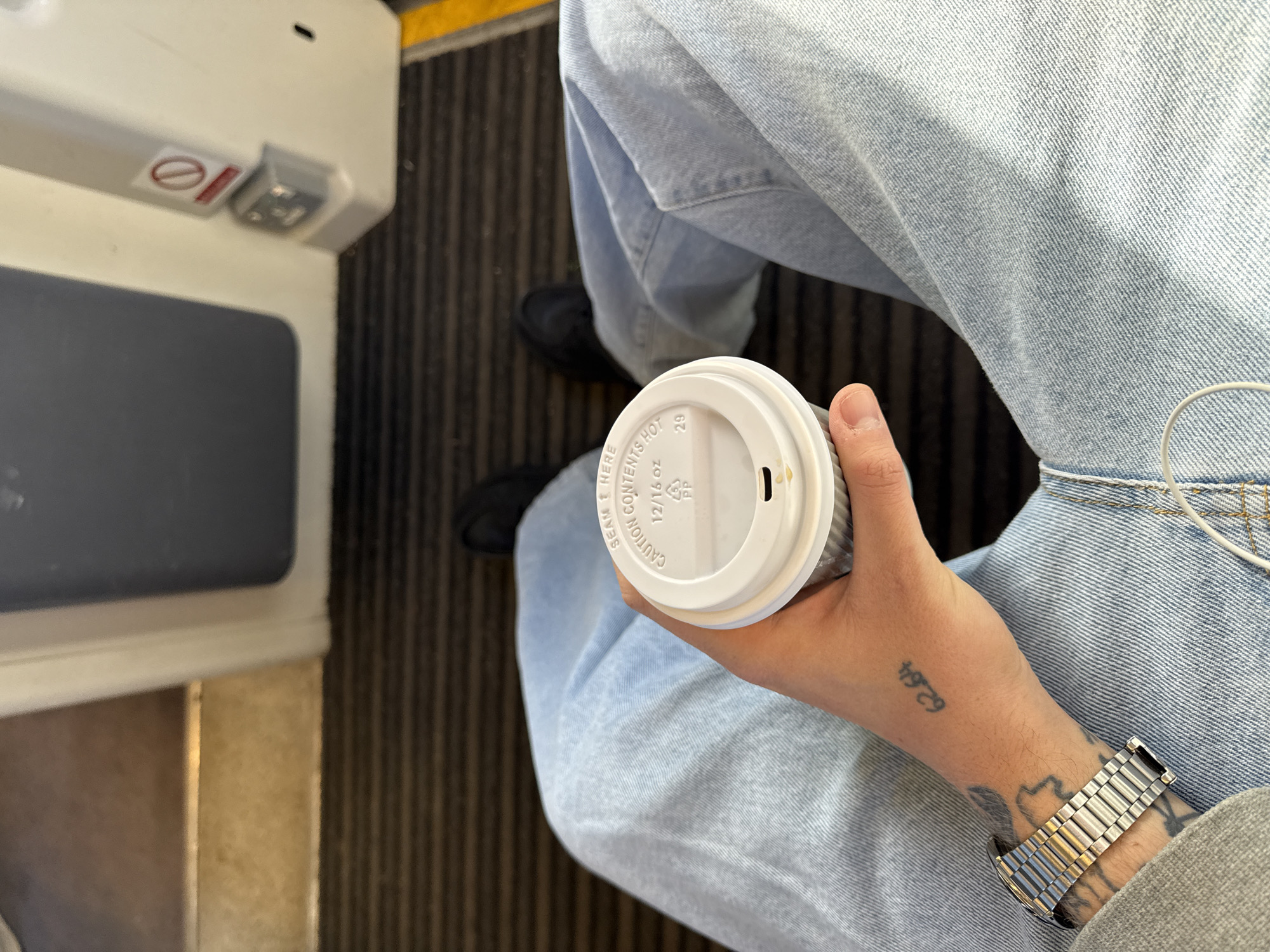The image captures a first-person perspective inside what appears to be a public transport vehicle, likely a train or bus, in North Hertfordshire, United Kingdom. The primary subject is a person's lower body and left arm, dressed in light-wash blue jeans and black shoes, sitting down.
The person's left hand holds a white disposable coffee cup with a white plastic lid. Visible text on the lid includes "CAUTION CONTENTS HOT," "12/16 OZ," a "PP" recycling symbol, and the number "29." The person's left wrist wears a metallic watch, and tattoos are visible on their arm and hand, one of which appears to be "62.64" or "62'64". White headphones or earbuds are also draped near the person's lap.
To the left, a light-colored interior paneling is visible, featuring a dark grey seat or floor panel. On the white panel, there's a red "no" symbol (a circle with a diagonal line through it), typically indicating a prohibition like "no smoking" or "do not block," next to a small grey button or switch. The floor features a striped, dark-colored carpet or mat. A yellow strip runs along the edge of the floor in the upper left background, commonly seen in train interiors or platform edges. The activity depicts someone traveling while holding a hot beverage. The lighting suggests daytime.