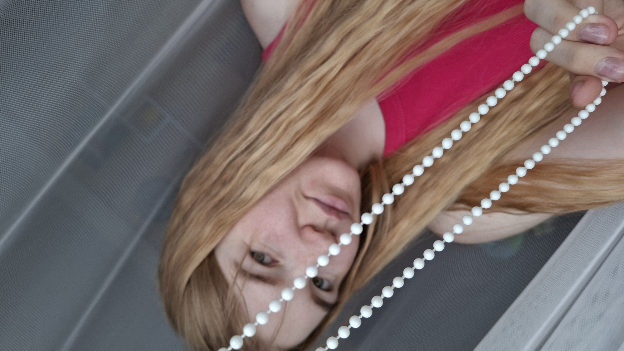 The image shows a young woman with long, wavy, light brown or blonde hair, wearing a bright pink top. The photograph is oriented sideways, with the woman positioned as if lying down and looking upwards towards the camera. Her face, featuring fair skin, visible freckles, and light-colored eyes, displays a soft, slightly smiling expression.
She is holding a white beaded chain with her right hand, which has light pink nail polish. The chain, composed of many small, round white beads, extends across the lower central portion of the frame.
The background consists primarily of a grey, semi-transparent, mesh-like or perforated surface, suggesting a window screen or part of a blind system. Through this mesh, faint and blurry colored elements are vaguely discernible in the upper left, indicating objects or a view beyond the screen. The scene appears to be set indoors during daytime, with natural light illuminating the subject. The overall mood conveyed is relaxed and pleasant. There is no visible text in the image. This scene is located in Bolshoy Bukor, Russia.