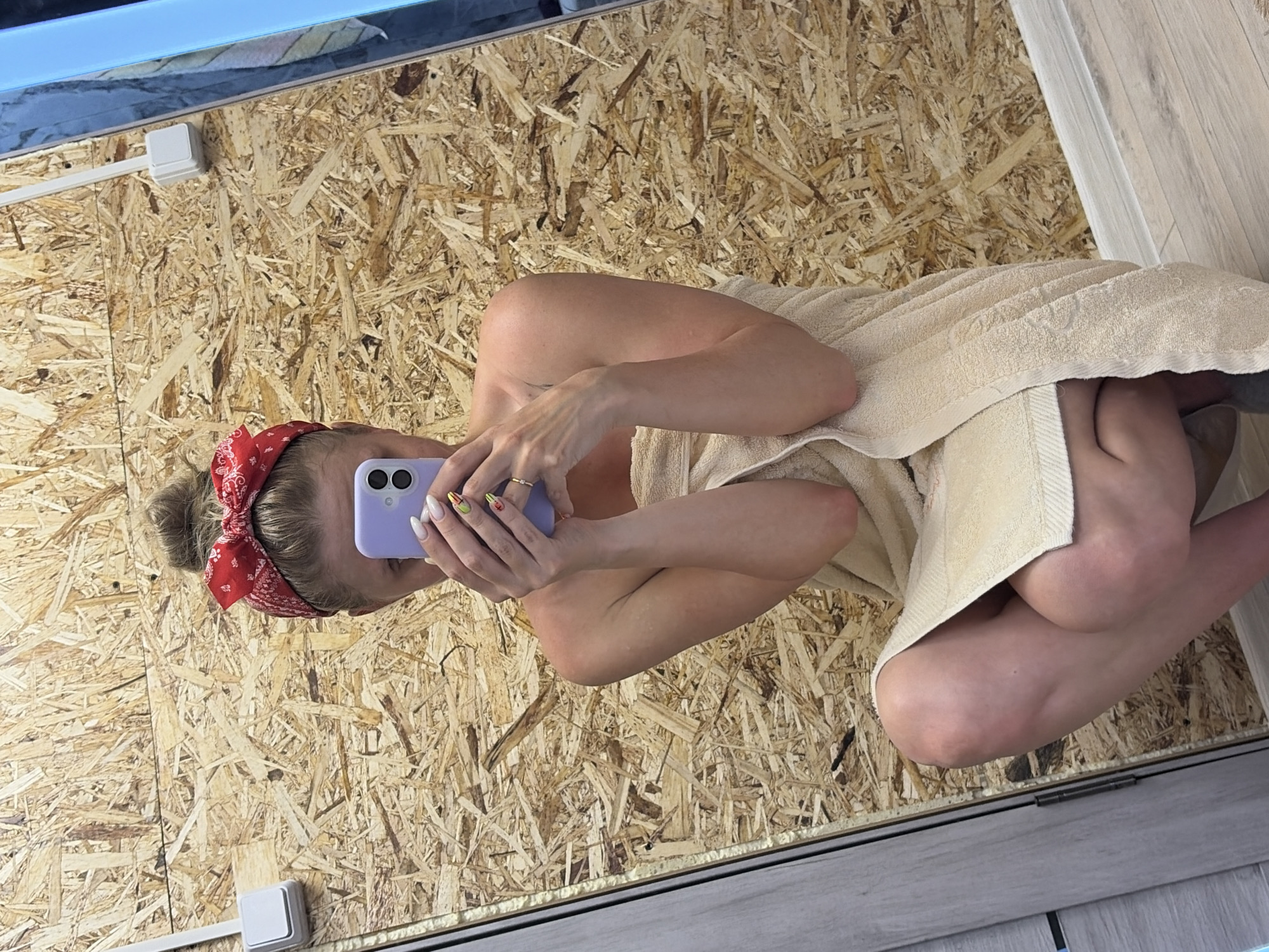 A woman with a red bandana in her hair takes a selfie in a mirror while sitting with a towel wrapped around her body. The background consists of a wall made of oriented strand board. Part of the ceiling and a section of a door frame are visible. The photograph appears to be taken indoors. The city of Chaikovsky, Russia is not discernible in the image.