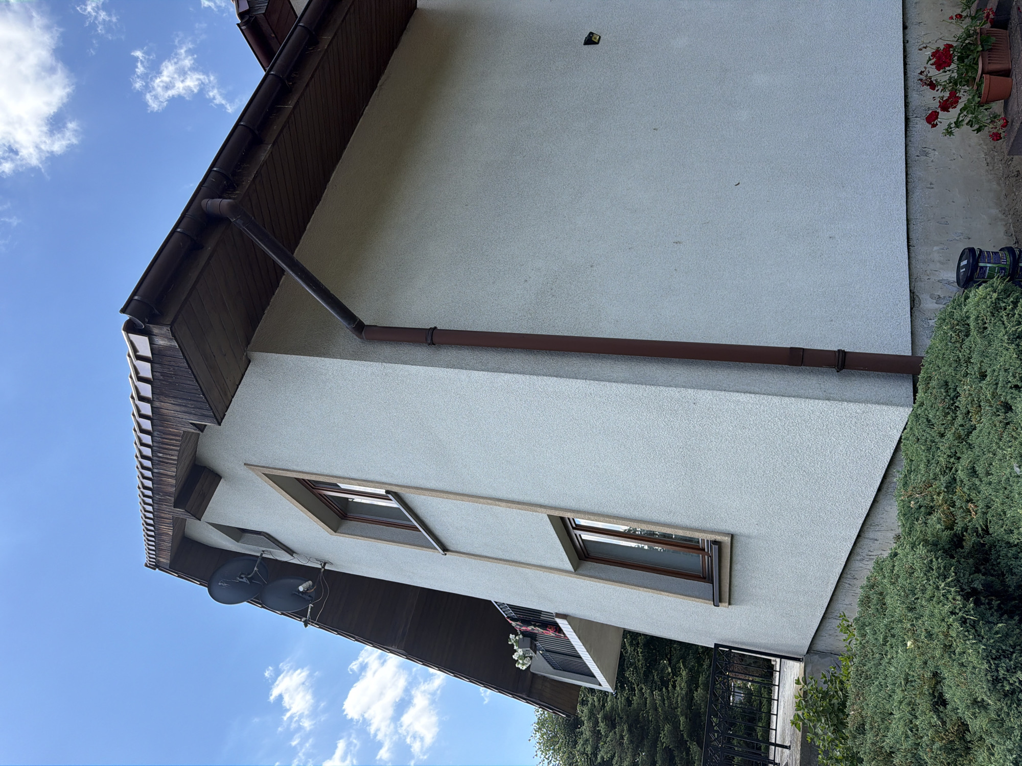 This is a side exterior view of a light-colored, stucco-sided house with a brown, wooden-shingled roof in Strzyżowice, Poland. The house has a sloping roofline, and two dark satellite dishes are mounted on the upper portion of the exterior wall near the eaves. There are two windows with dark brown frames visible on this side of the house. A dark brown gutter system is installed along the roofline, with a downspout running vertically down the side of the house. A small balcony with a black metal railing is visible on the left side of the house, with white flowers in a planter. Green bushes and trees are in the foreground and on the left side of the image. Red flowers in pots are visible at the base of the house on the right. The sky is blue with scattered white clouds.