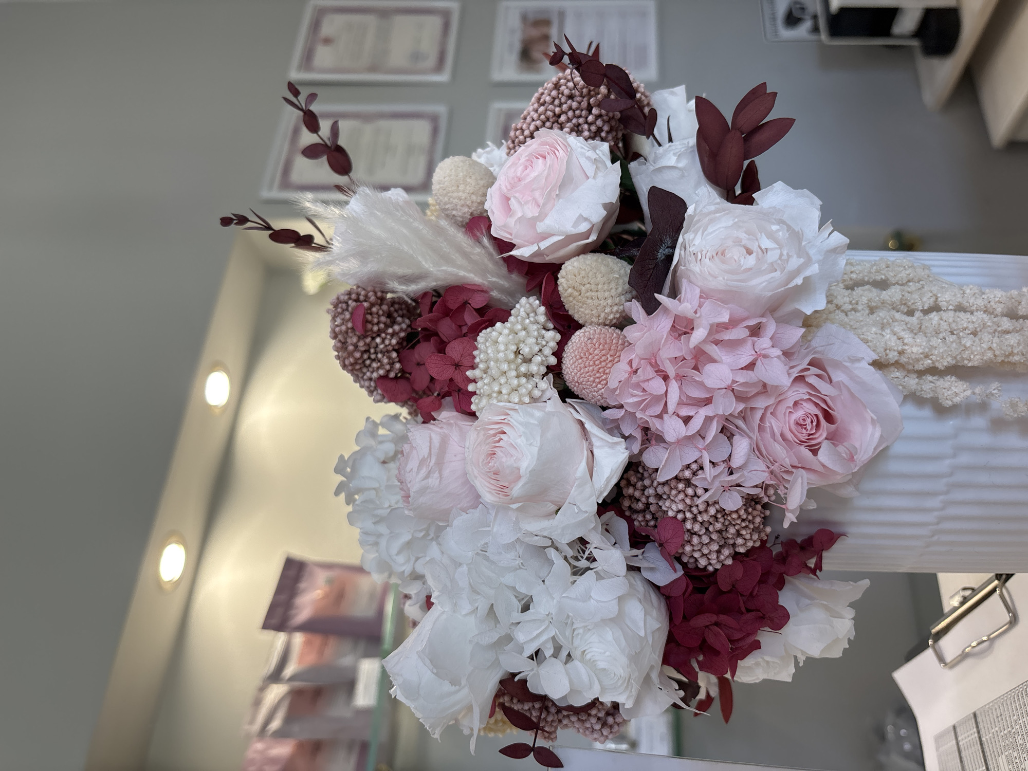 A close-up, eye-level shot captures a lush bouquet of preserved flowers, predominantly in shades of pink, white, and deep burgundy. The arrangement is composed of various textures, featuring delicate rose blooms, fluffy pampas grass, and clusters of small, round blossoms. Dark red, dried leaves add a dramatic contrast. The bouquet is placed in a white, textured vase, and a sliver of what appears to be a white pleated surface is visible to the right. The background is a soft gray wall with what looks like framed certificates or awards hanging in the upper portion. Two warm-toned lights are visible on the left side of the image, illuminating a section of the wall and a display of items that are out of focus. The overall impression is one of a carefully crafted floral arrangement, possibly for decoration or a special occasion, presented in a well-lit indoor setting. There is no visible text within the frame.