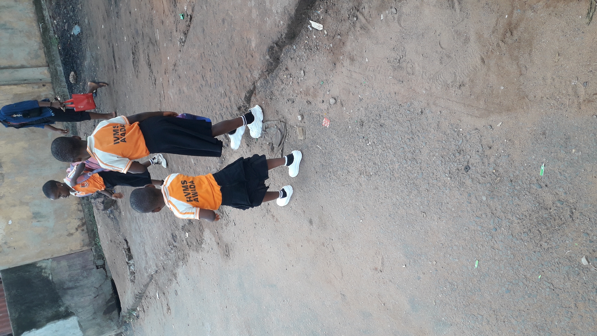 On an unpaved road in Onitsha, Nigeria, a group of children wearing orange school uniforms with "IVMS AWADA" printed on the back are walking. They are wearing black skirts or shorts and white shoes. There are four children visible, with a possible fifth partially obscured. The environment is a mix of dirt and cracked pavement, with a building or structure visible on the left side of the frame.