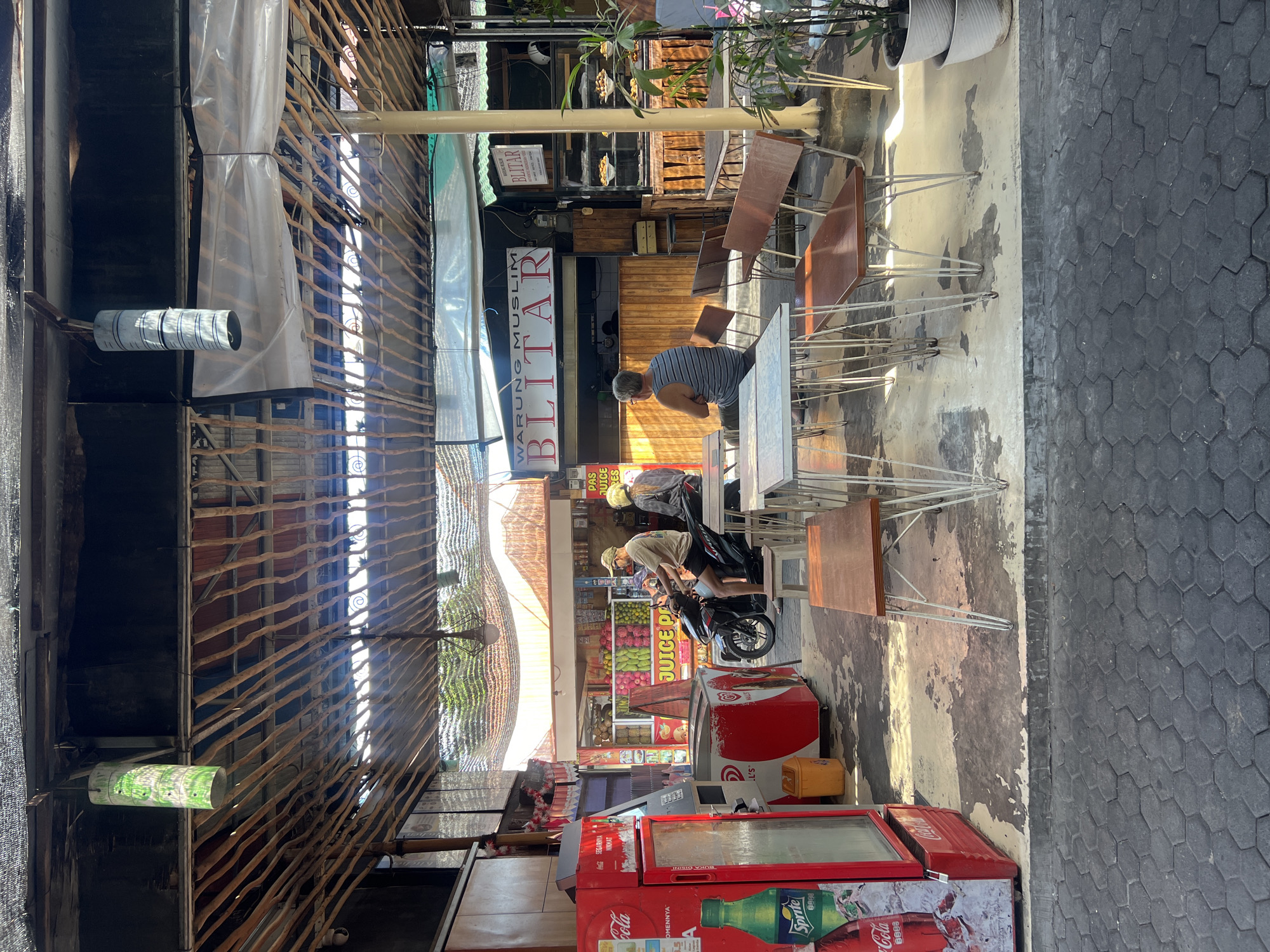 In Kuta, Indonesia, an open-air food stall with a bamboo-slat roof is depicted. The stall features a red Coca-Cola refrigerator on the left, stocked with ice and beverages. Adjacent is a white and red ice cream freezer. Several tables and benches made of metal and wood are arranged in front of the stall, some occupied by patrons. A man in a striped shirt sits at a table facing away from the camera. Further into the stall, a counter displays food items, and a sign reads "WARUNG MUSLIM BLITAR." Two individuals on a motorcycle are visible near the counter. The ground is paved with hexagonal stones. Hanging lanterns and various plants contribute to the environment.