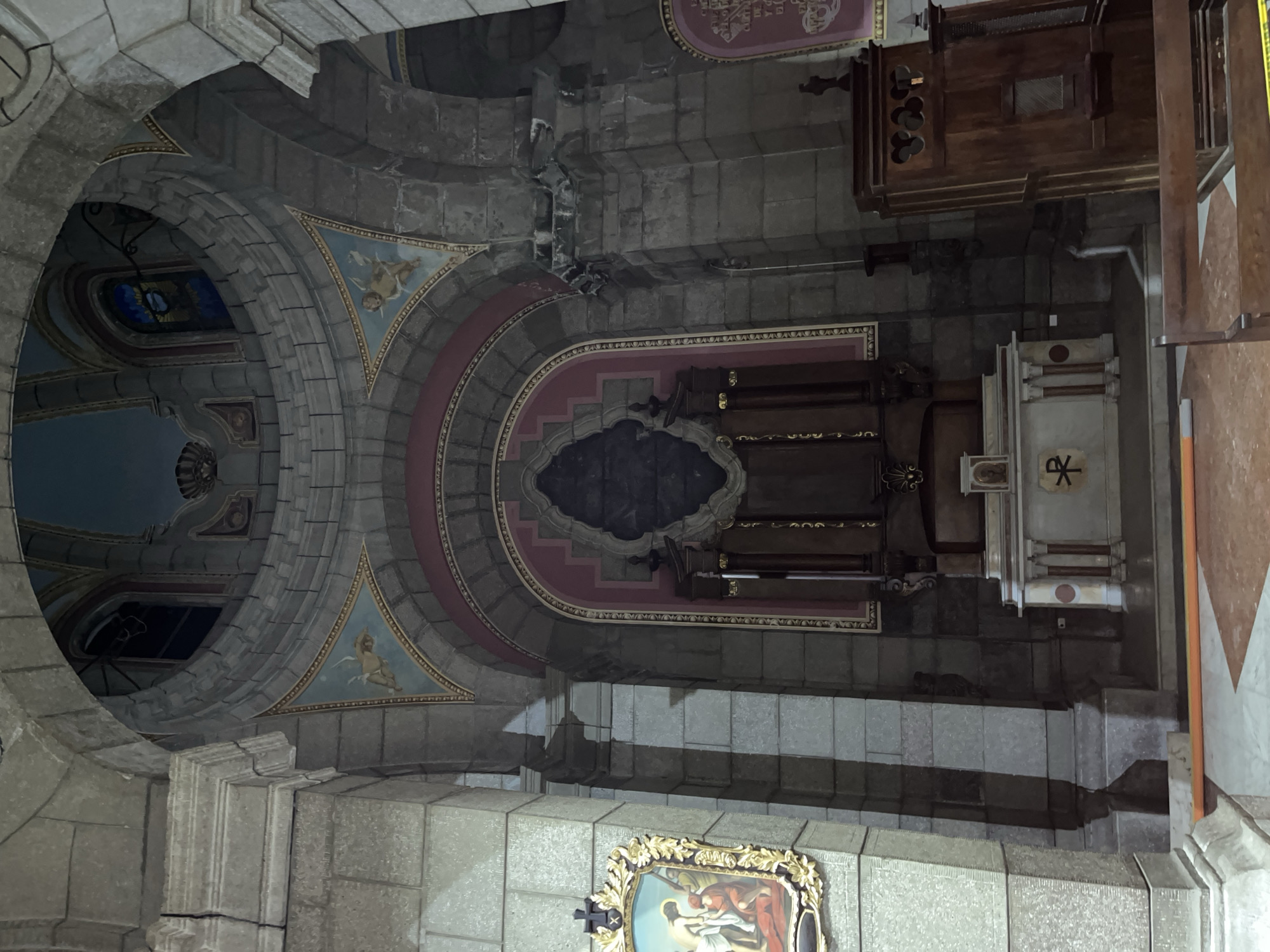 The image shows the interior of a church, likely a cathedral, in Mérida, Venezuela. The architecture features stone columns and arches, with a domed ceiling adorned with frescoes depicting angelic figures. In the foreground, an altar is visible, complete with a cross and religious symbols. To the left, a painting of a religious scene hangs on the wall. To the right, a wooden confessional booth is positioned. The lighting suggests an indoor environment, with natural light filtering in from the windows in the dome. The overall impression is one of solemnity and historical grandeur, characteristic of a place of worship.