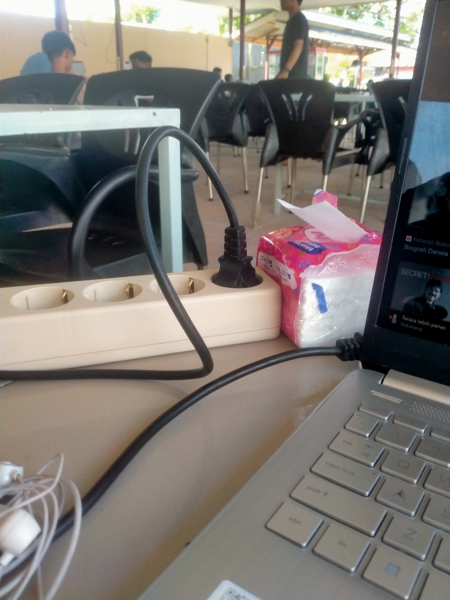 The image shows a laptop computer resting on a table in an outdoor setting, possibly a cafe or a similar public space, in an area identified as "Unknown, Unknown". The laptop's screen is partially visible, displaying a video or website. Next to the laptop, there is a box of tissues. The laptop is connected to a power strip via a black power cable. The power strip is plugged into a wall outlet. A tangled set of white earphones is also visible near the laptop.
Several people are seated at tables in the background, suggesting a public or social environment. The tables and chairs appear to be part of an outdoor seating arrangement under a roof or awning. The lighting suggests daylight conditions.