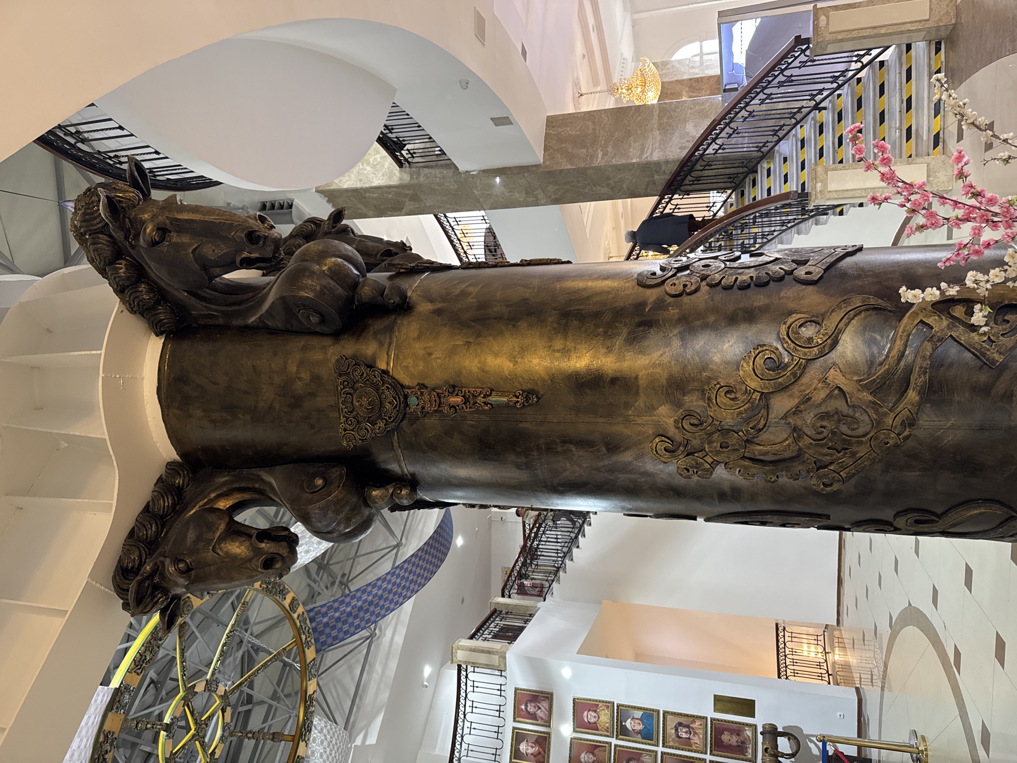 The image captures the interior of a multi-story building in Ulaanbaatar, Mongolia, showcasing a large, cylindrical art installation with horse head sculptures at both ends. The bronze-colored piece is adorned with intricate, embossed patterns along its surface. It is positioned vertically in the center of the building's atrium, visible against a backdrop of staircases, marble walls, and tiled floors.
On the upper floors, staircases with black railings are visible, one featuring yellow and black caution strips. A person wearing a dark coat is walking up one of the staircases.
The lower floors feature a display of framed portraits lining the walls, and a wheelchair ramp is visible near the bottom of the frame. The interior design combines modern and traditional elements, with the art installation being a focal point. The environment appears well-lit and spacious.