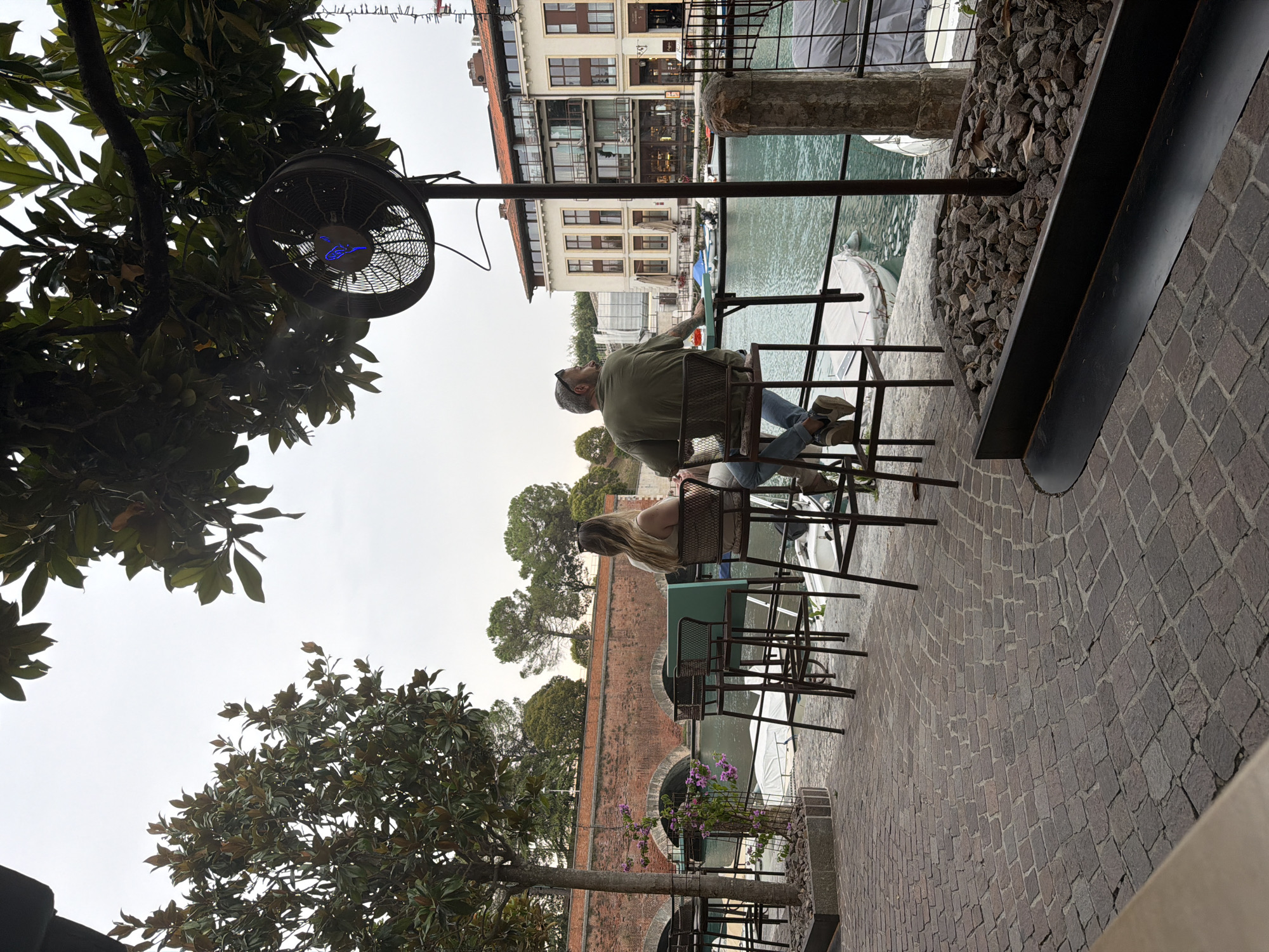 A couple sits at a table on an outdoor patio overlooking a canal in Peschiera del Garda, Italy. The woman, with long blonde hair, wears a light-colored top and jeans, and the man, with a shaved head and beard, is dressed in a green t-shirt and jeans. They are seated on high chairs, facing away from the camera and towards the water. The canal is lined with boats and buildings with terracotta roofs. A brick bridge spans the canal in the background. The patio is paved with cobblestones and features a metal railing and a large outdoor fan hanging from a tree. The scene is set on an overcast day.