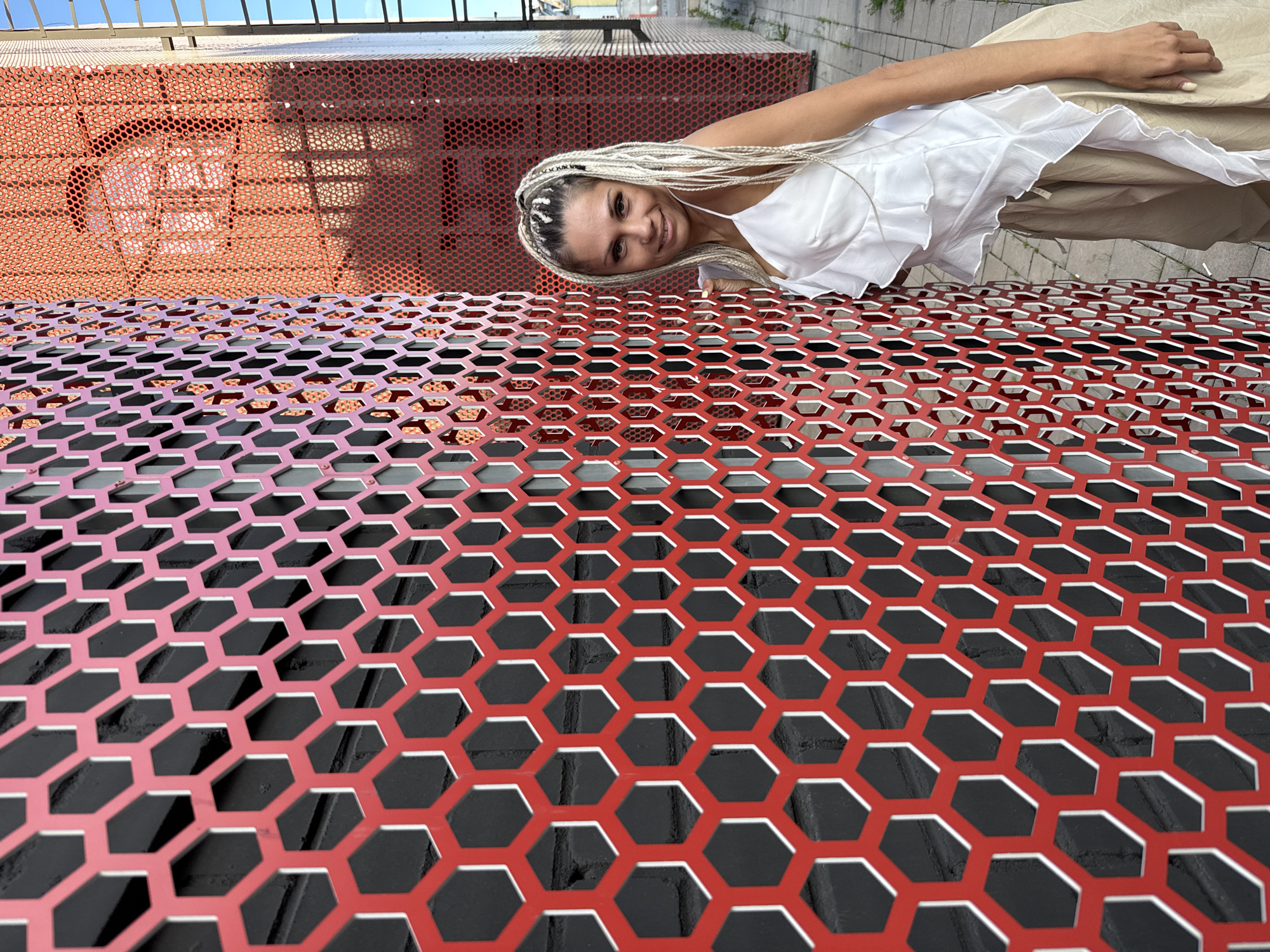 A woman with long, blonde braided hair leans against a red, hexagonal metal screen in an urban environment. She is wearing a white ruffled top and tan pants. The background shows a brick wall behind the screen, and further back, a red building with a similar perforated facade. The ground is paved with gray stones. The scene appears to be outdoors during the daytime, with shadows indicating sunlight. The location is specified as Esbol Rural District, Kazakhstan.