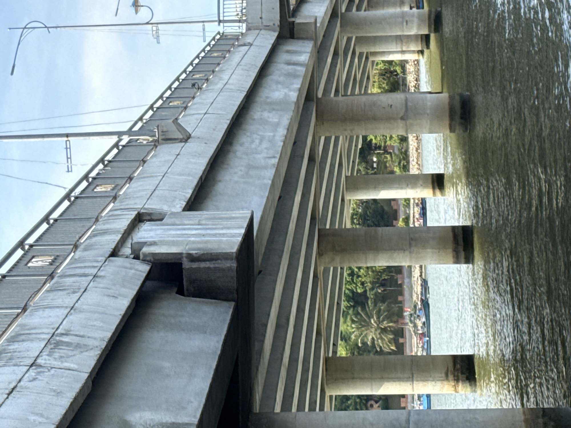 The image shows a concrete bridge spanning over a body of water, likely a river or bay. The bridge is supported by numerous concrete pillars. The water appears choppy with ripples. In the background, across the water, there is a shoreline with lush green trees, including palm trees. Several boats are visible on the water near the shore. The sky is overcast with some clouds. The scene appears to be in a coastal or riverside urban area.