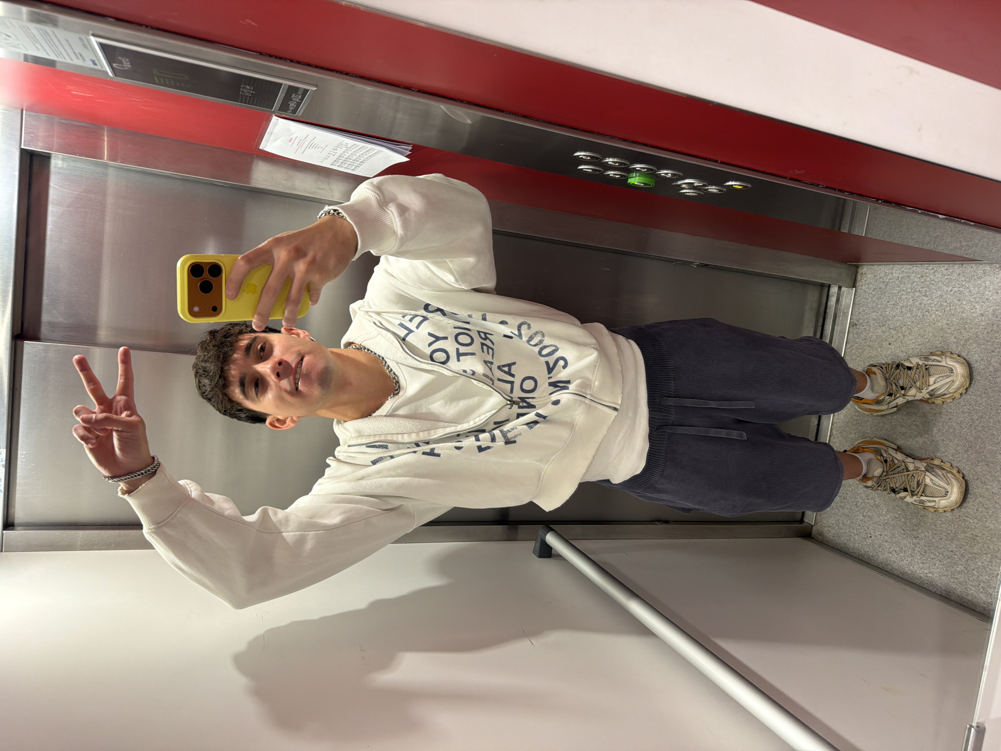 The image shows a young adult male taking a selfie in an elevator. He's wearing a white sweatshirt with a design, blue shorts, and athletic sneakers. He is holding a yellow smartphone and making a peace sign with his left hand. The elevator has red and white panels at the top, a control panel with buttons, and a metal interior. The floor is gray and speckled. The location is in Sofia, Bulgaria.
