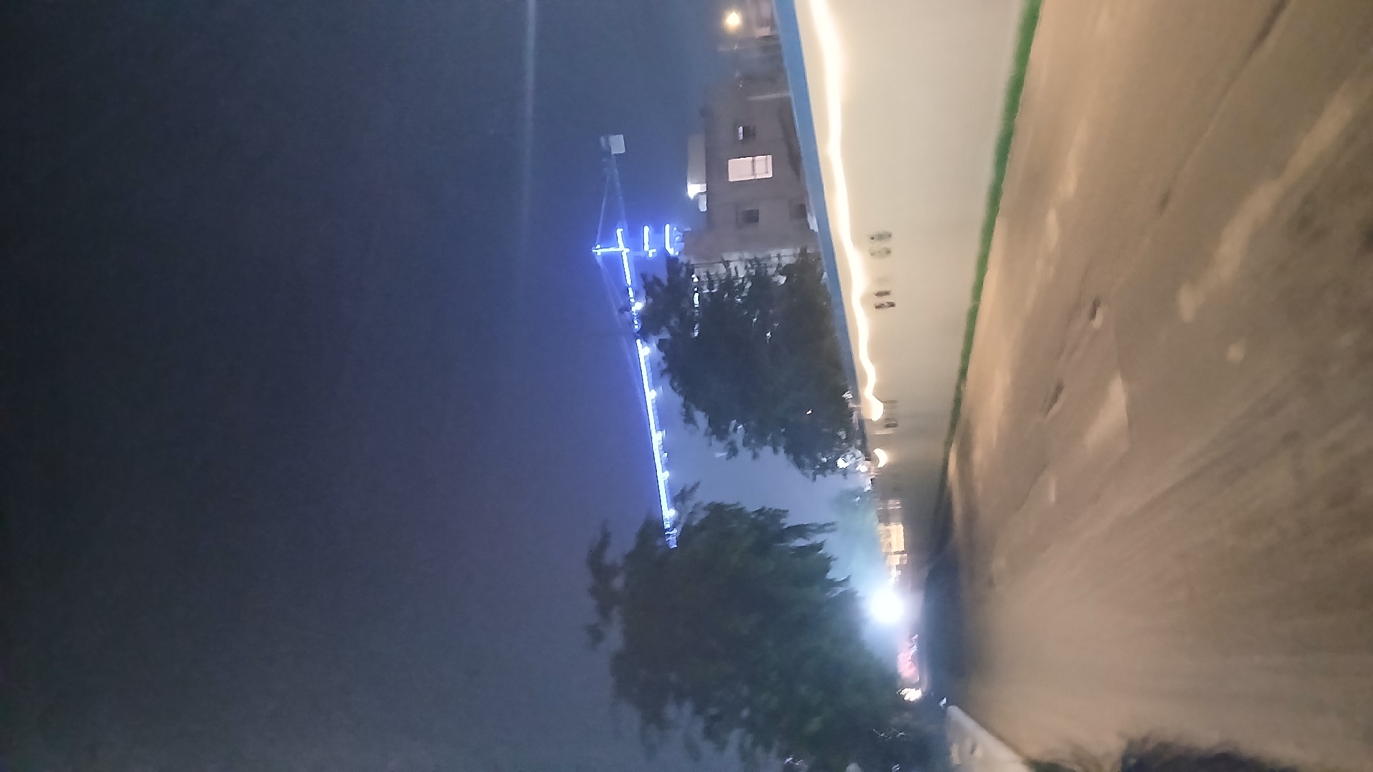 The image depicts a nighttime scene on a road in the City of Medan, Indonesia. A white, illuminated wall runs along the right side of the road, adorned with decorative lights. Beyond the wall, a building is visible, with windows lit from within. In the background, a large construction crane, illuminated with blue lights, stands against the dark sky. Two trees with dense foliage are situated on the left side of the road, partially obscuring the view of the building and crane. The road itself appears to be under construction or in disrepair, with visible patches and uneven surfaces. The overall atmosphere is dark, with artificial lighting from the wall, building, and crane illuminating the scene. There are no people or vehicles clearly visible in the image.