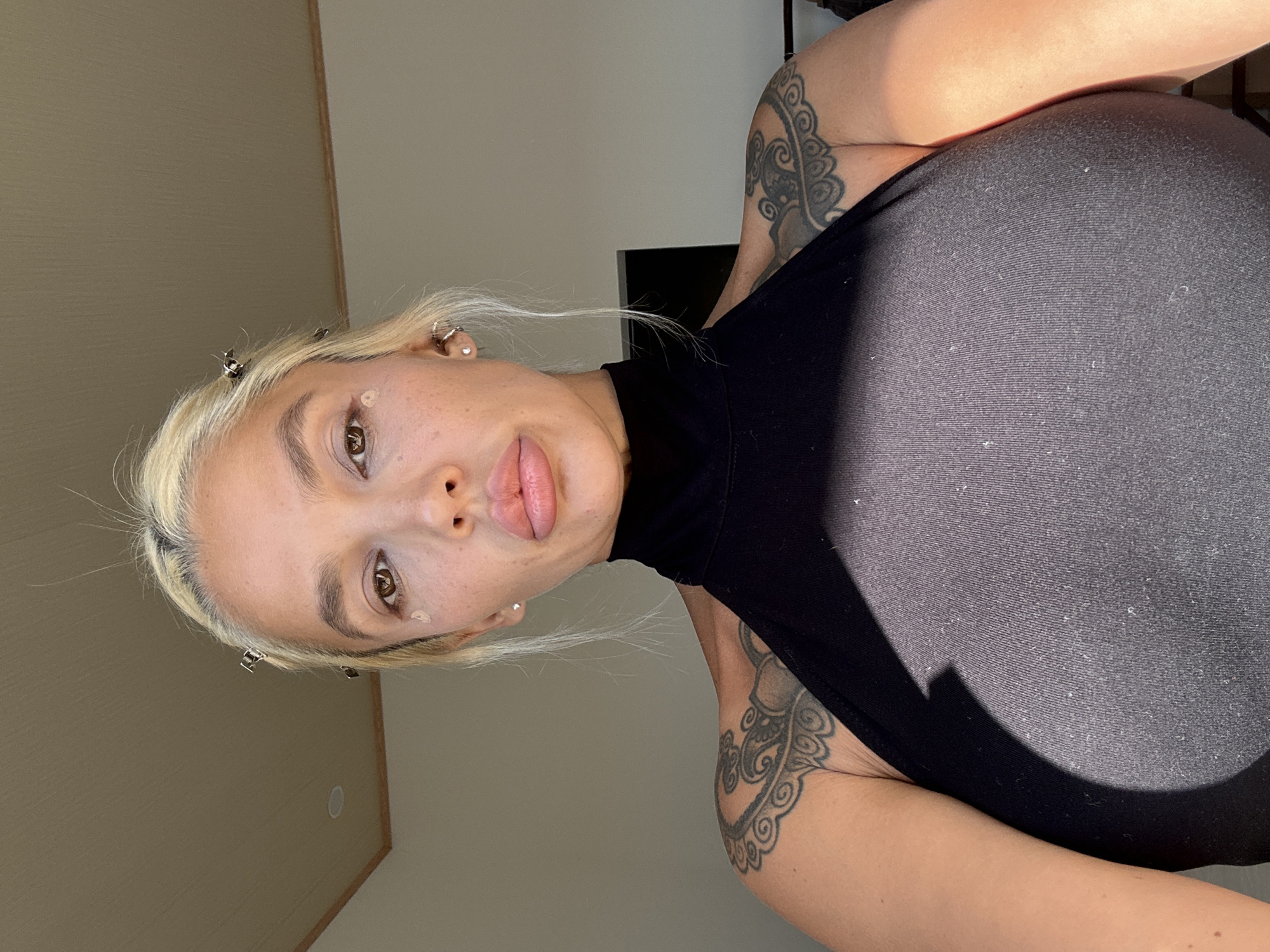 A light-skinned woman with blonde hair is angled in an indoor scene. She is wearing a black top. She has tattoos on her upper arms. Her hair is held back with clips, and she has small objects stuck to her face. The location is unknown, and there's no indication it's in Kuta Selatan, Indonesia.