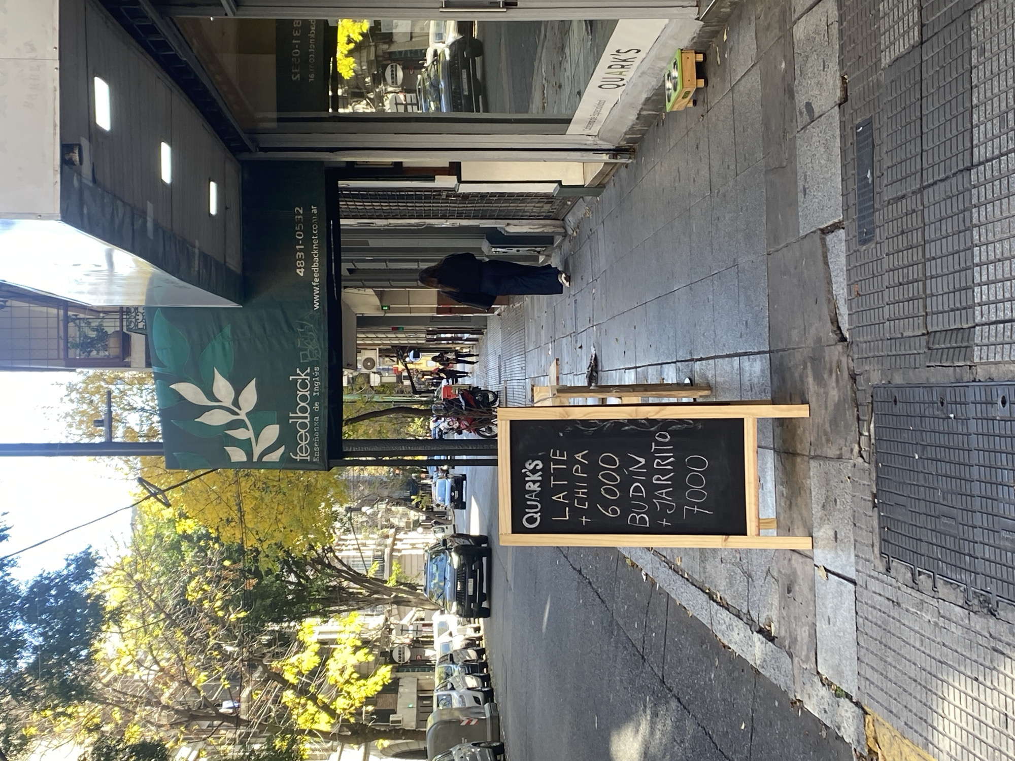 The photograph depicts a street scene in Buenos Aires, Argentina, likely during the day, as indicated by the natural lighting. The camera is positioned at an eye-level perspective, capturing a sidewalk and a street.
A black sign board with white lettering stands on the sidewalk advertising "Quark's Latte + Chipa 6000" and "Budin + Jarrito 7000". The sign is placed on the sidewalk, near a curb.
Adjacent to the sidewalk is a street with cars parked along its side. The street leads into the distance, where other vehicles and pedestrians can be seen. Trees with yellow leaves line the street, suggesting it is autumn.
To the left of the sign, a banner labeled "feedback Enseñanza de Inglés" is visible. A phone number and website are also on the sign.
A person is seen walking near the storefront. The scene presents a typical urban environment with commercial establishments, street furniture, and the daily activities of city life.