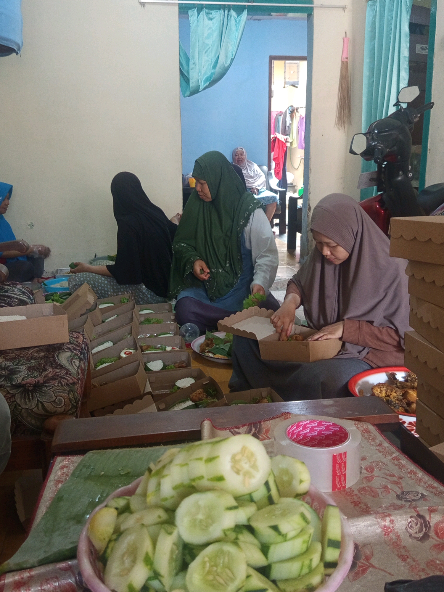 In Mulyorejo, Indonesia, a group of women are engaged in the preparation of food boxes. The scene is indoors, with a motorcycle visible in the background. Several women, dressed in hijabs, are seated on the floor surrounded by numerous cardboard food boxes, many of which are filled with rice, vegetables, and what appears to be fried chicken. One woman in a green hijab is actively placing food into a box, while another in a brown hijab is also arranging food items. A third woman in a black hijab is partially visible. In the foreground, a bowl of sliced cucumbers sits on a table, next to a roll of clear tape with a red label. The environment suggests a communal activity, likely for distribution or an event.