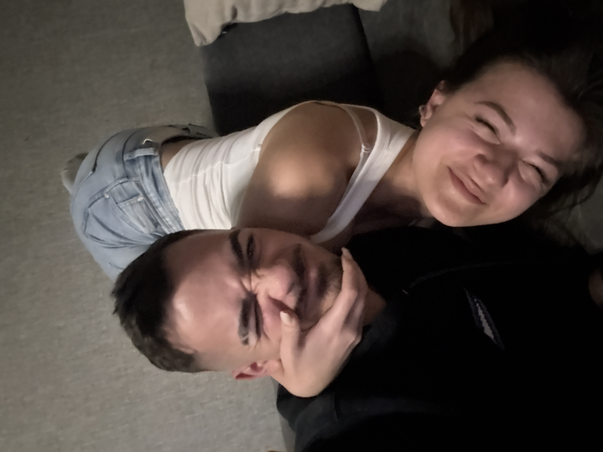 The image shows two people in an indoor setting, possibly a home. A woman is on top of a man and has her hand on his face, seemingly playfully choking him. The woman is wearing a white tank top and blue jeans. The man is wearing a black hooded sweatshirt. The background includes a gray couch and carpeted floor. There is no indication of the location being in Sofia, Bulgaria.