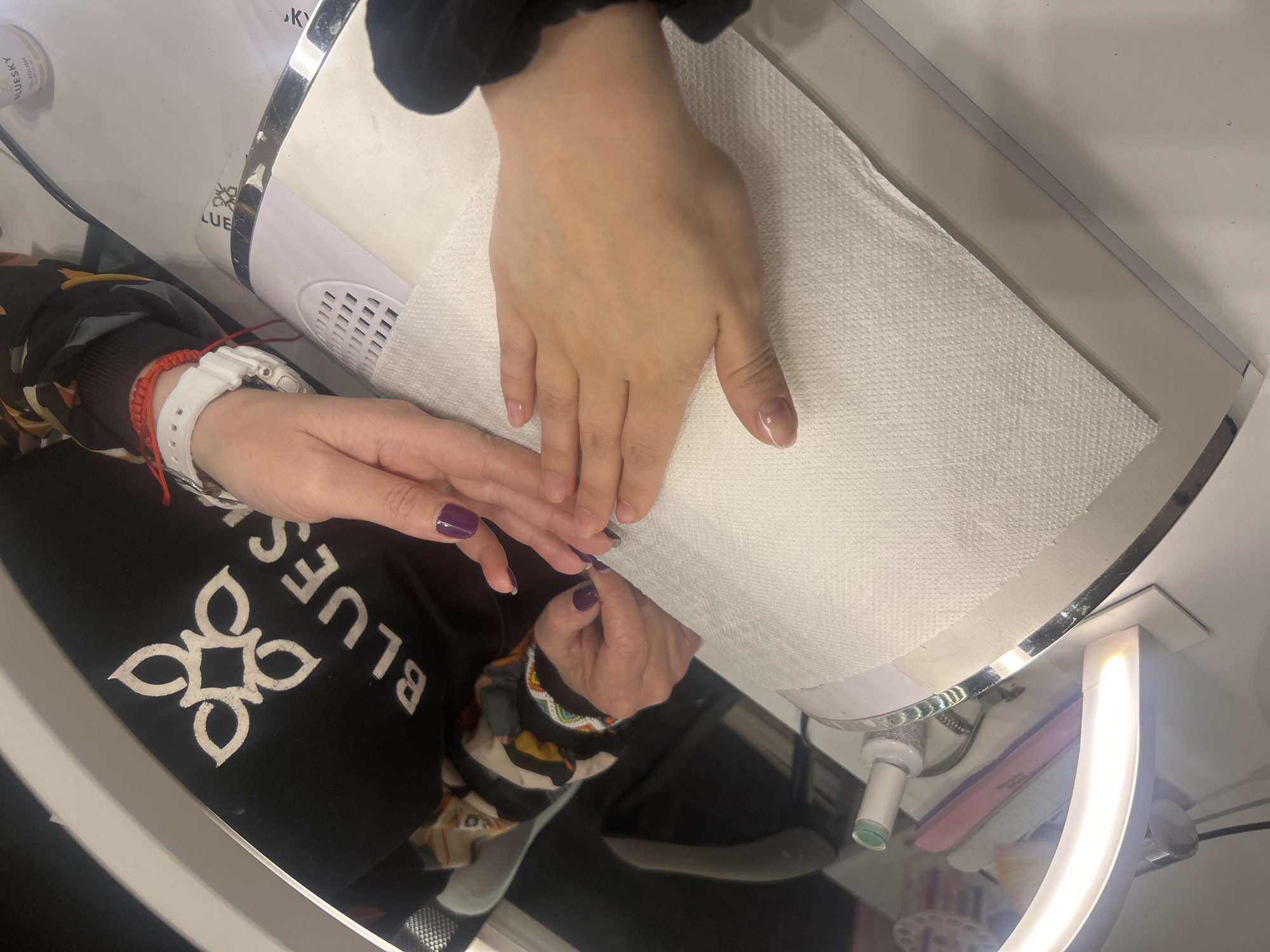 Two hands are visible in the scene. One hand is resting palm down on a white paper towel inside of a nail lamp. The nails on this hand appear to be bare. The other hand is to the left. The nails on this hand are painted a dark purple. The person whose hands are in the scene is wearing a black sweater with a white design and the word "BLUES" printed on it. A white watch and red string bracelet are visible on the wrist of the hand with the purple nails. The scene appears to be taking place in a nail salon. The location is unknown.