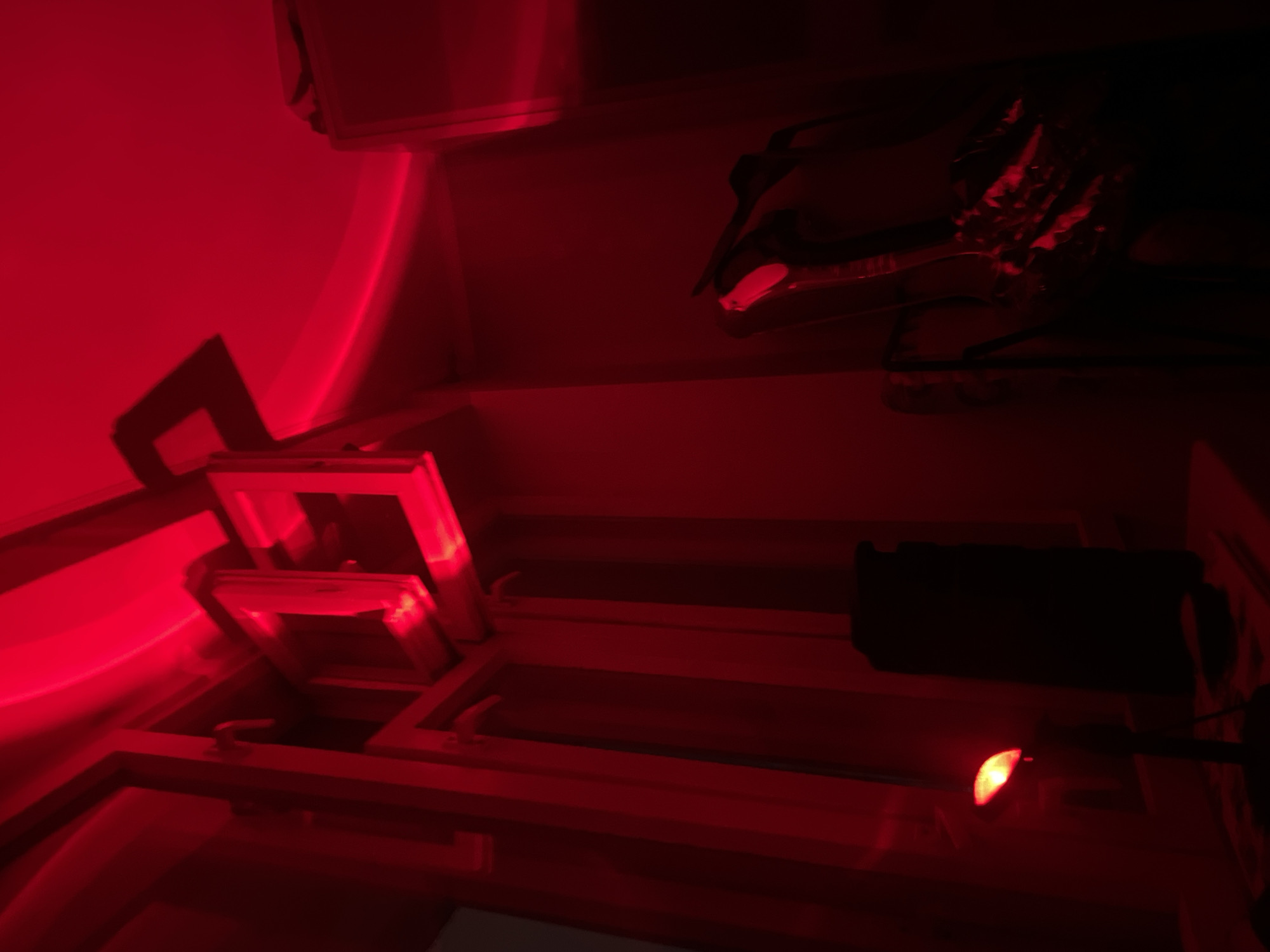The image displays an indoor scene illuminated entirely by a deep red light, strongly suggesting a photographic darkroom environment. In the lower right foreground, a small, bright red light source, likely a safelight, is visible, attached to a dark, rectangular object that could be part of a piece of equipment or a handle.
Towards the mid-left, two prominent frame-like structures are arranged, possibly developing tanks or trays, reflecting the intense red light on their surfaces. The room features light-colored walls and curved architectural elements, uniformly aglow with the red illumination, creating a sense of depth and structure. In the upper right section, a dark, complex object with metallic and somewhat jagged features is partially visible, also reflecting the red light, implying it is another piece of darkroom equipment or a specialized tool.
No human presence or direct activity is depicted, but the setup of the room and the specialized lighting clearly indicate an environment prepared for or actively used for photographic processing. No visible text is present in the image.