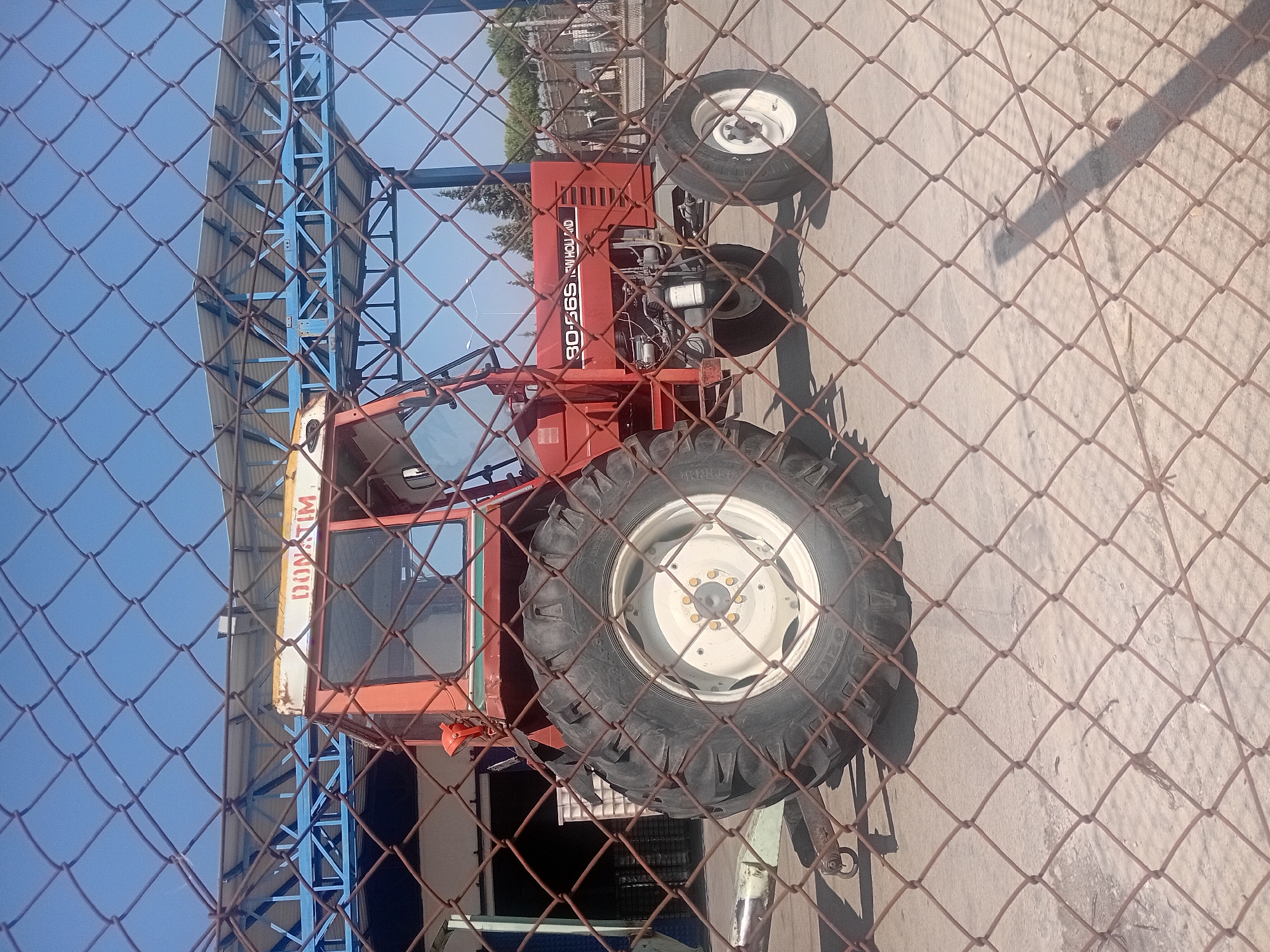 The image shows a red New Holland tractor, model 80-66S, parked behind a chain-link fence. The tractor is orange-red with a white roof on the cabin. Text on the cabin reads "DONATIM" and the model designation "80-66S NEW HOLLAND" is visible on the side of the tractor. The tractor has large, rugged tires and smaller front wheels. The scene appears to be outdoors, possibly a yard or industrial area, with a blue metal structure visible behind the tractor, suggesting a covered area or shelter. The ground is paved concrete. The sky is clear and blue. The image is taken from a low angle, looking through the diamond-shaped openings of the rusty chain-link fence. The presence of the tractor and the industrial-looking shelter suggests a location related to agriculture or machinery storage.
