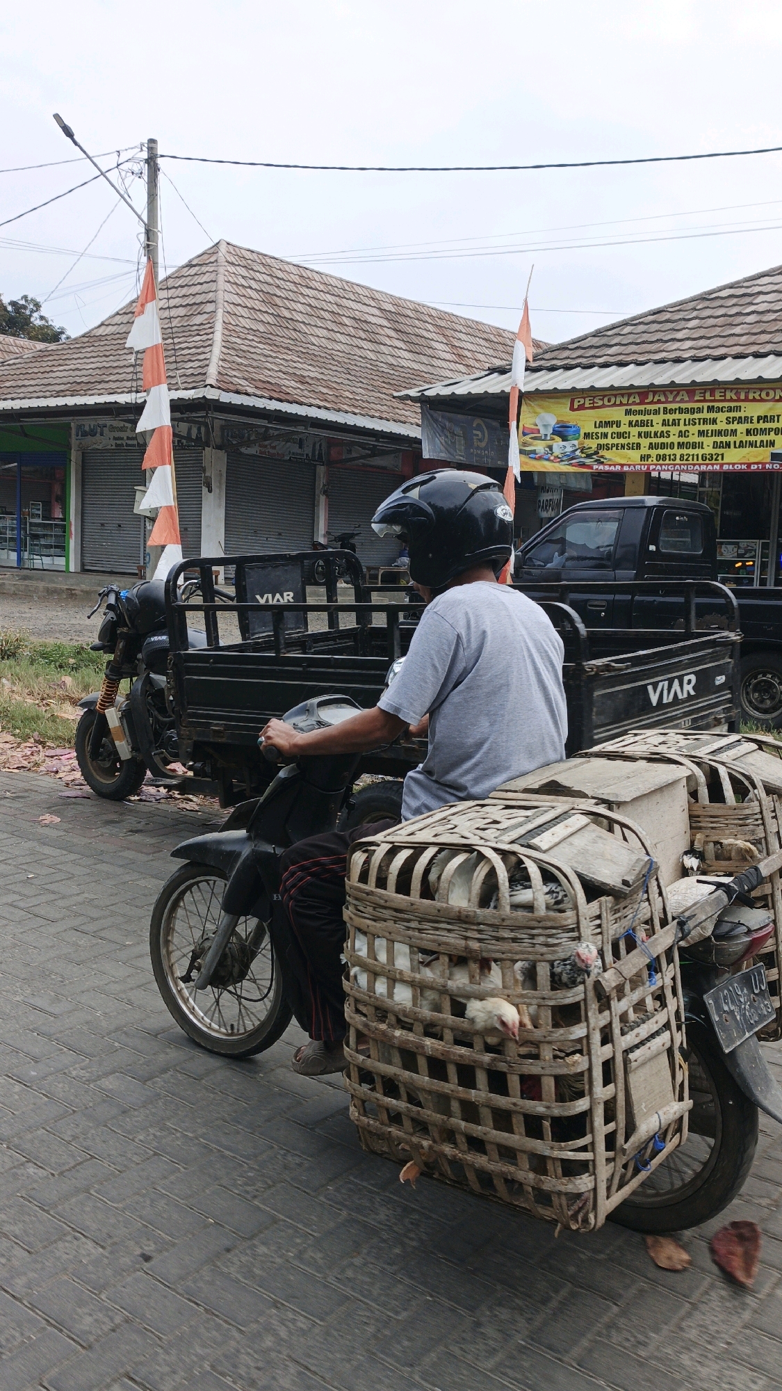 In Pangandaran, Indonesia, a person wearing a gray t-shirt and black helmet rides a black motorcycle with a cargo carrier. The motorcycle is loaded with two large woven bamboo baskets filled with live chickens. The motorcycle has a license plate that reads "GJ 9244 UJ". In the background, there are buildings with red tile roofs and shops with closed shutters. One shop has a yellow banner with text in Indonesian. A black truck is parked behind the motorcycle. The scene takes place on a paved road.
