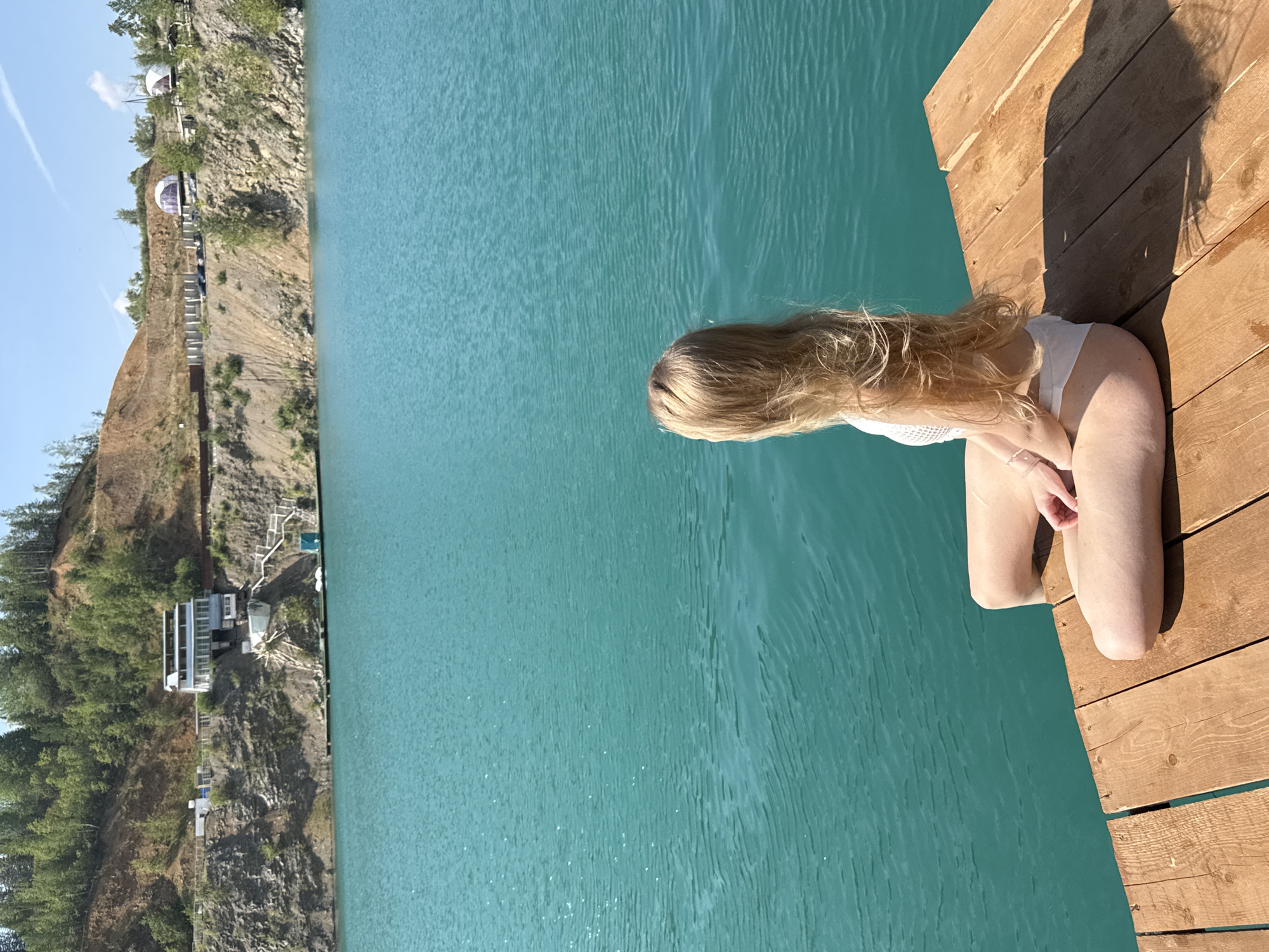A young woman with long blonde hair, wearing a white bikini, sits cross-legged on a wooden dock facing away from the camera, towards a large body of turquoise water. In the background, the steep, eroded banks of a quarry rise up, topped with trees. A multi-story building with a flat roof and many windows is visible on the upper part of the bank, along with a set of stairs leading down towards the water. Further along the bank, two geodesic domes are visible. The sky is blue with some wispy clouds. This location is Karyer Izvestnyak, Russia.