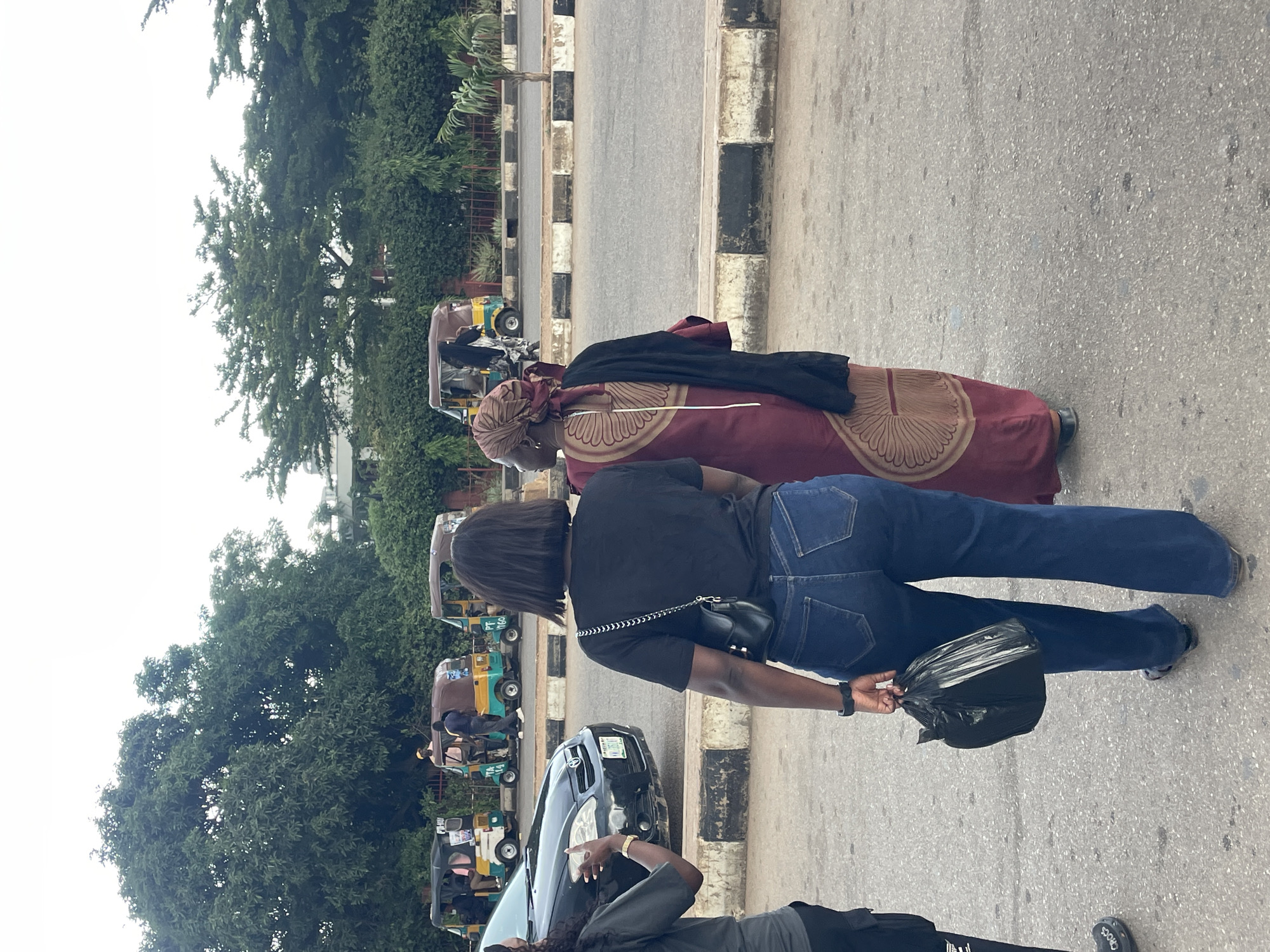 In Jos, Nigeria, two women stand on a paved road beside a black sedan. The woman on the left, facing away from the camera, wears a dark t-shirt and blue jeans. She holds a black plastic bag in her right hand and a black purse on her left shoulder. The woman on the right, also facing away, wears a maroon dress with a circular pattern and a headscarf, with a black shawl draped over her shoulders. In the background, several auto-rickshaws are parked along the roadside. Dense green trees and foliage line the far side of the road, with a pale, overcast sky above.